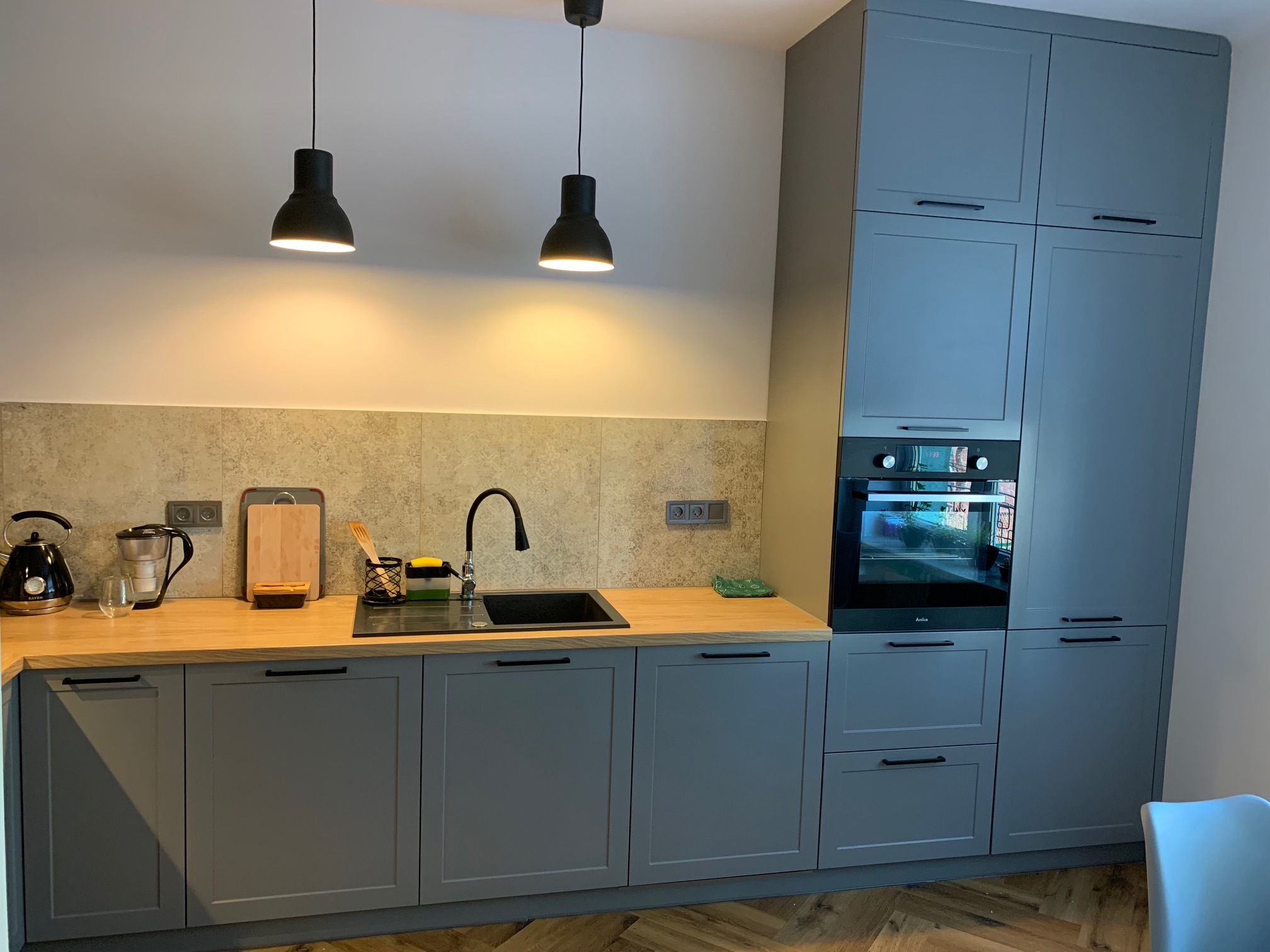 Szare meble kuchenne z drewnianym blatem, czarnym zlewozmywakiem i dwiema wiszącymi lampami w nowoczesnym stylu.