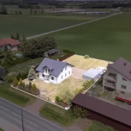 Nowoczesny, biały dom jednorodzinny z szarą dachówką, otoczony ogrodzeniem, widok z lotu ptaka na działkę z garażem i trawnikiem.