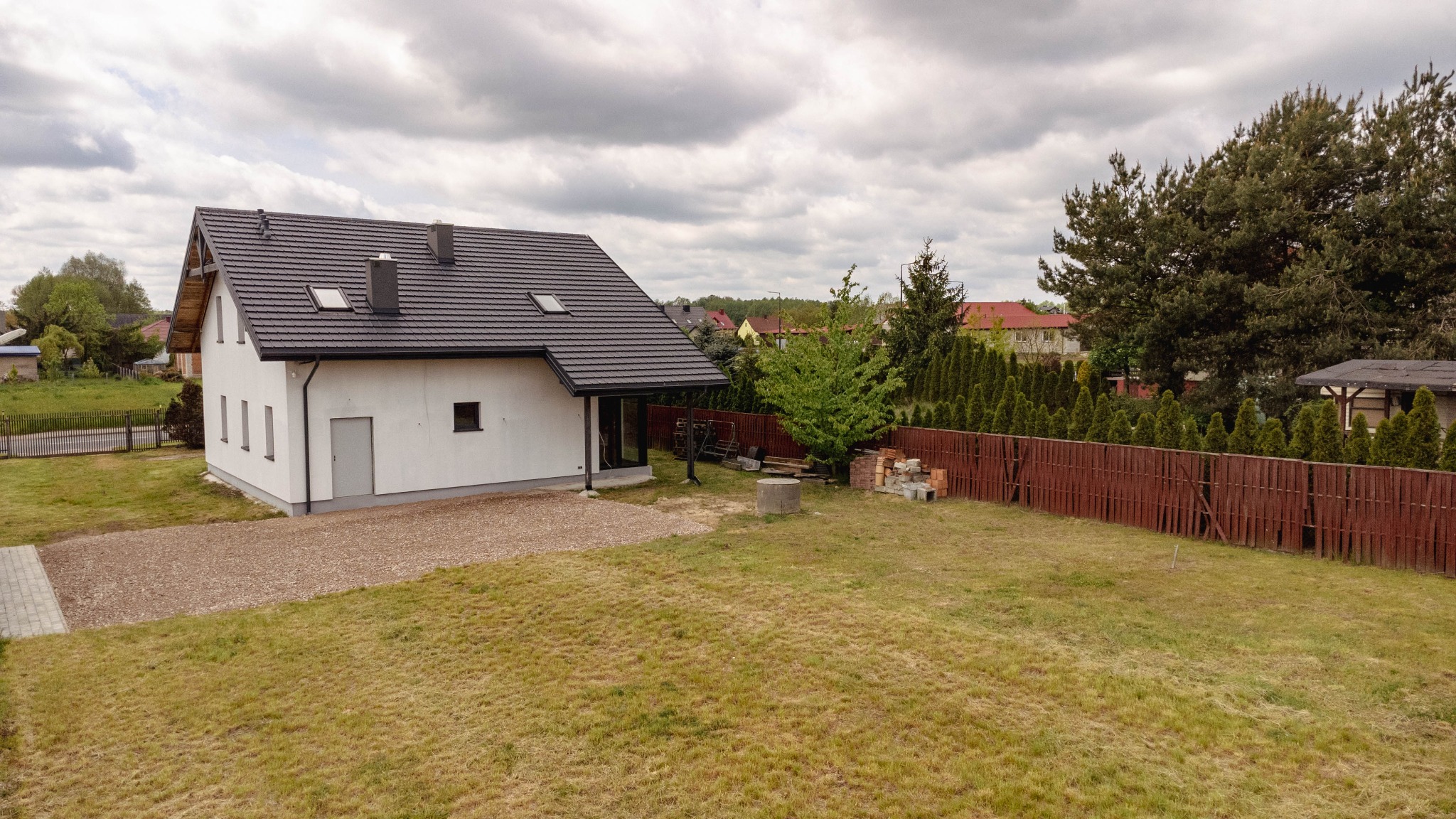 Nowoczesny, biały dom z ciemną dachówką, widok z zewnątrz na ogród z drewnianym ogrodzeniem i elementami budowlanymi