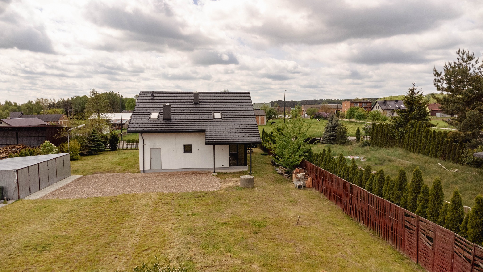 Nowoczesny, biały dom z ciemną dachówką i oknami dachowymi, widoczny z góry na tle pochmurnego nieba i zielonego ogrodu z drewnianym ogrodzeniem.