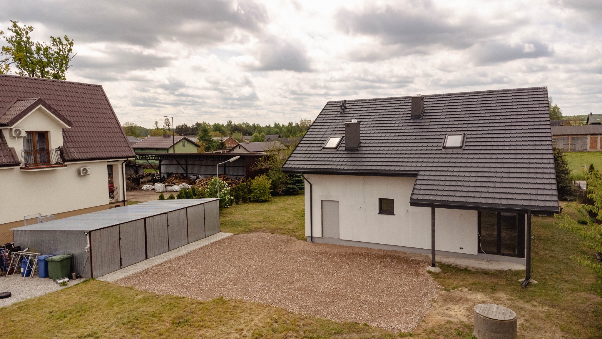 Nowoczesny, biały dom jednorodzinny z grafitowym dachem z dachówki, widok z góry, z przodu wysypany brązowy żwir, w tle inne domy i pochmurne niebo.