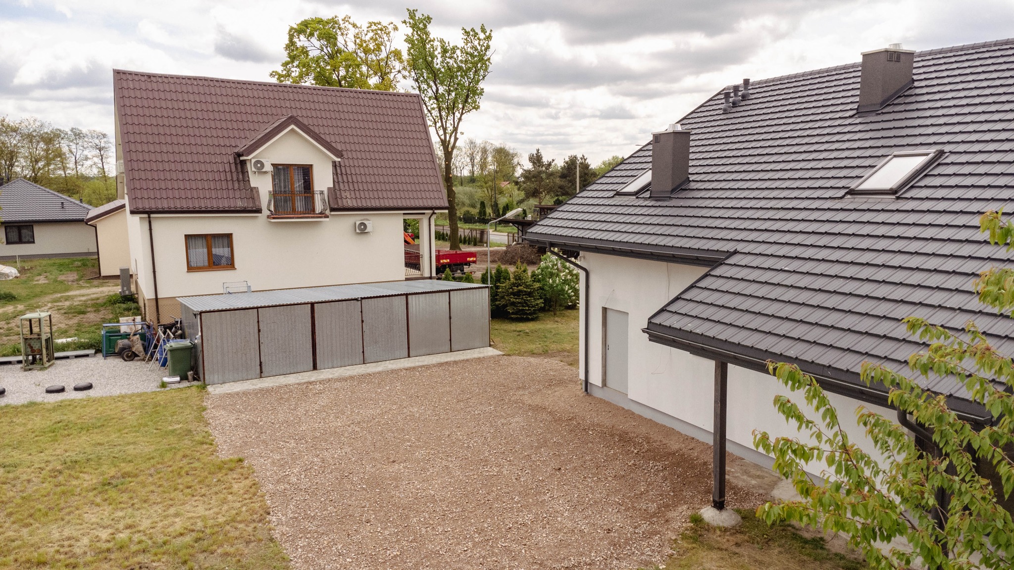 Dwa domy jednorodzinne z brązowymi i szarymi dachami, widok z góry na podwórze z żwirowym podłożem i blaszanym garażem.