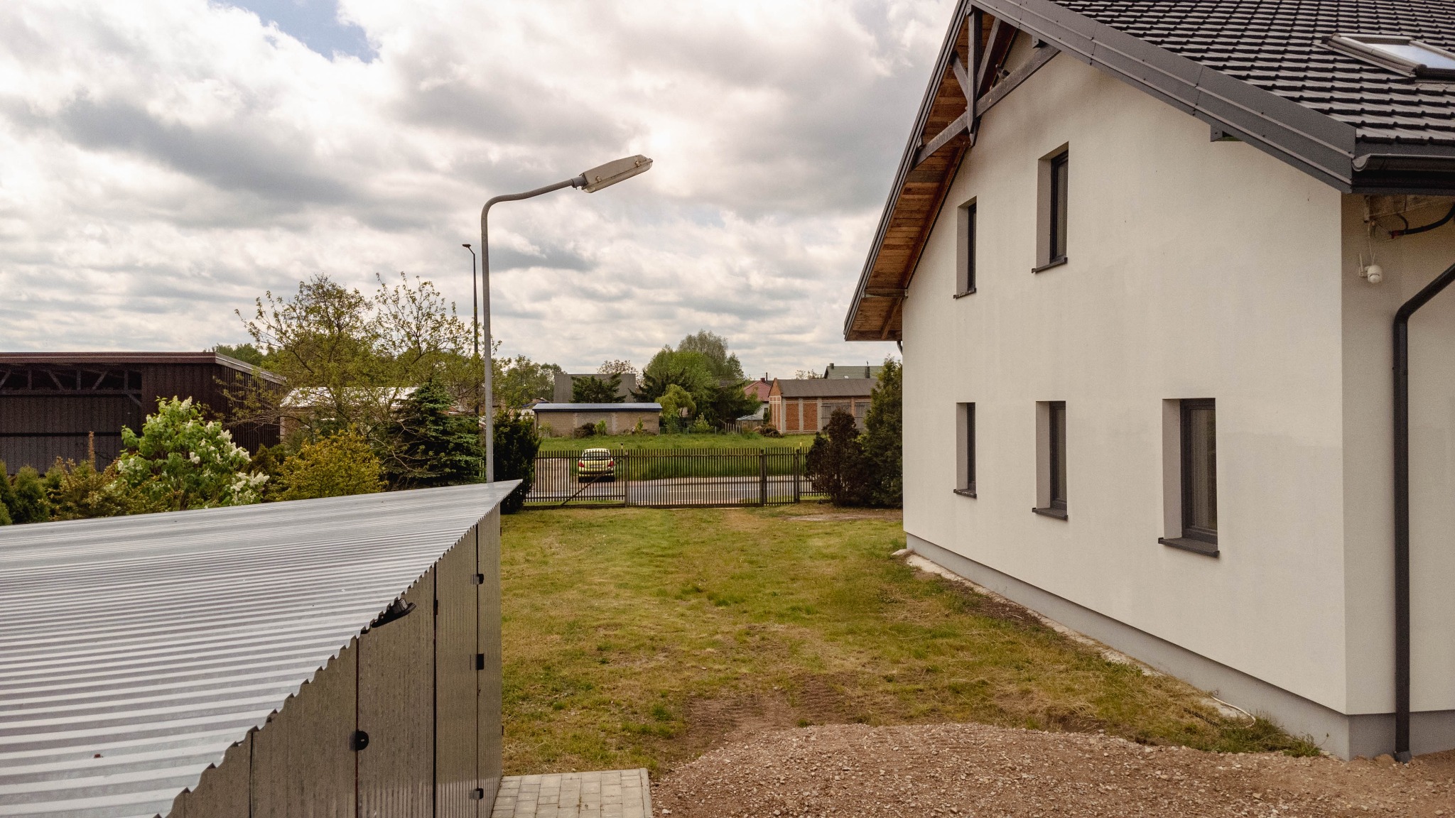 Nowoczesny dom jednorodzinny z białymi ścianami, czarną dachówką i trzema oknami na bocznej ścianie, widok z podwórka z metalowym garażem i lampą uliczną w tle.