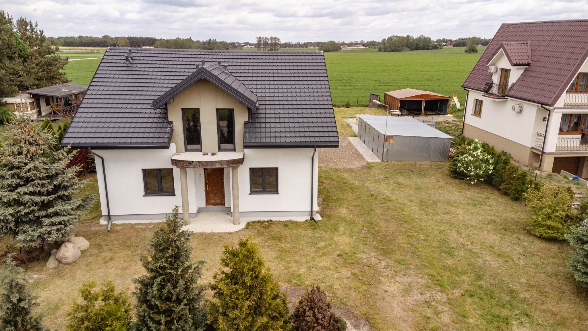 Nowoczesny, dwukondygnacyjny dom jednorodzinny z ciemną dachówką i białymi ścianami, w trakcie wykańczania elewacji, z widocznym ogrodem i garażem blaszanym w tle, sfotografowany z lotu ptaka.