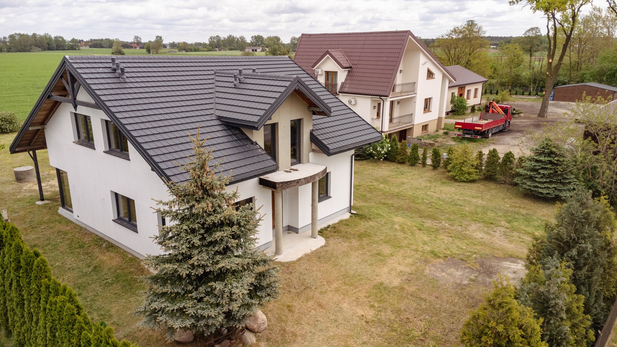 Nowoczesny, dwukondygnacyjny dom z ciemnoszarą dachówką i białymi ścianami, otoczony zielenią; w tle widoczny drugi dom z czerwoną dachówką i czerwona ciężarówka z dźwigiem.