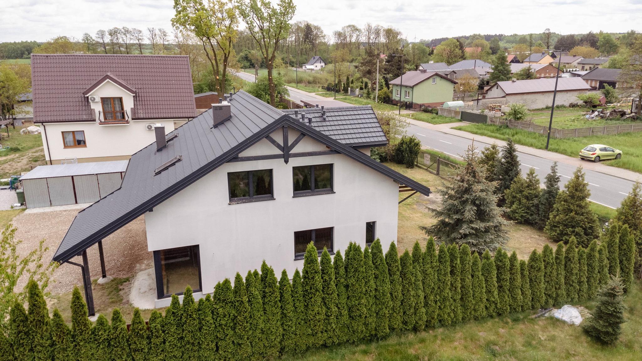 Nowoczesny, biały dom jednorodzinny z ciemnoszarą dachówką, widziany z góry, otoczony zielonym żywopłotem i innymi drzewami iglastymi, z widoczną drogą i zabudowaniami w tle.