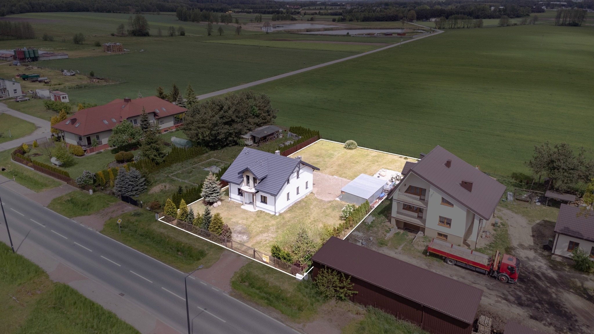 Nowoczesny, biały dom jednorodzinny z szarą dachówką, otoczony ogrodzeniem, widok z lotu ptaka na działkę z garażem i trawnikiem.