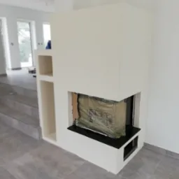 Nowoczesna obudowa kominkowa w trakcie montażu, z widocznym wkładem zabezpieczonym folią, jasne ściany i szare płytki podłogowe.