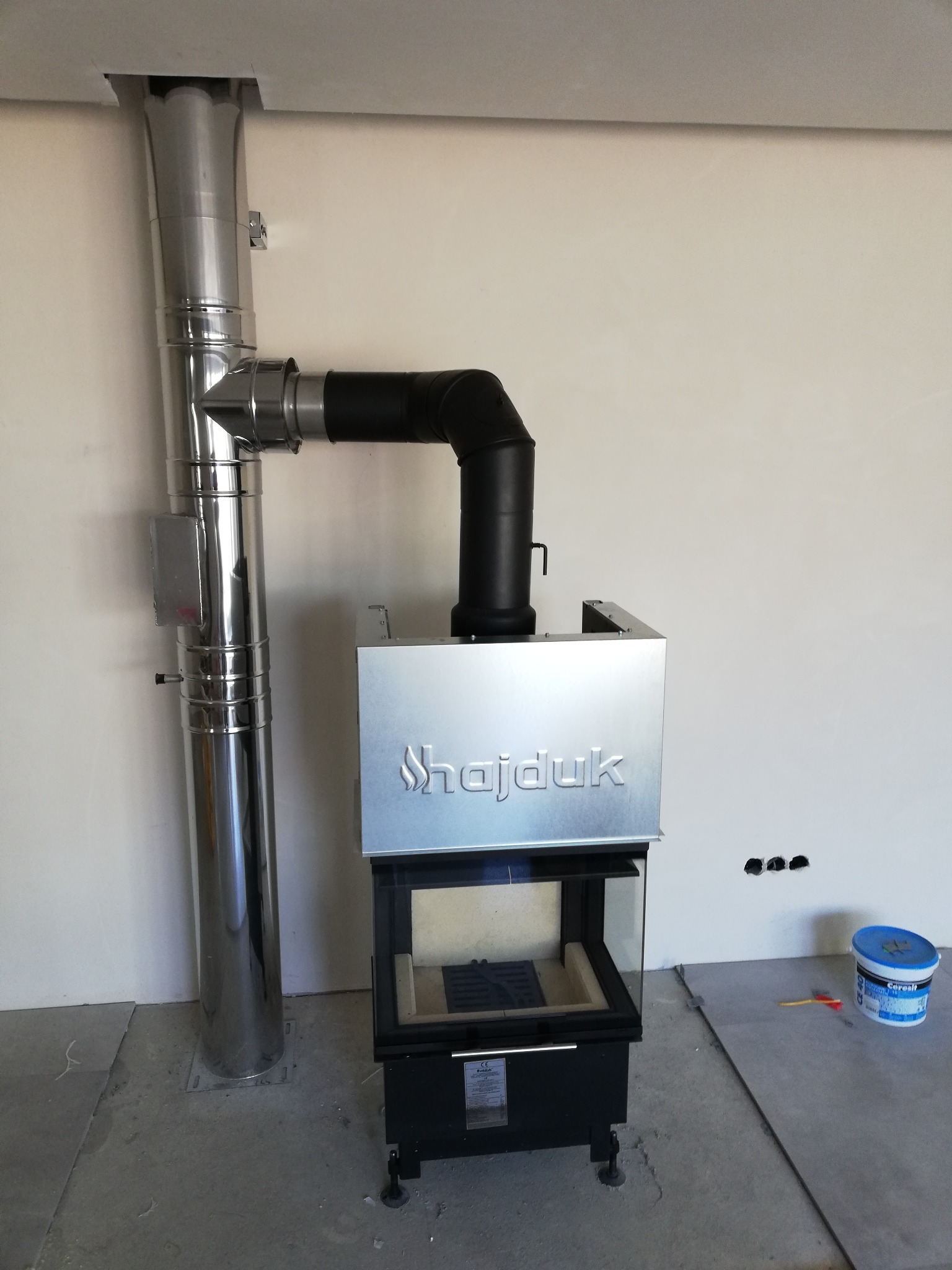 Nowoczesny wkład kominkowy marki Hajduk z podłączonym, błyszczącym kominem stalowym, w surowym wnętrzu w trakcie wykończenia.