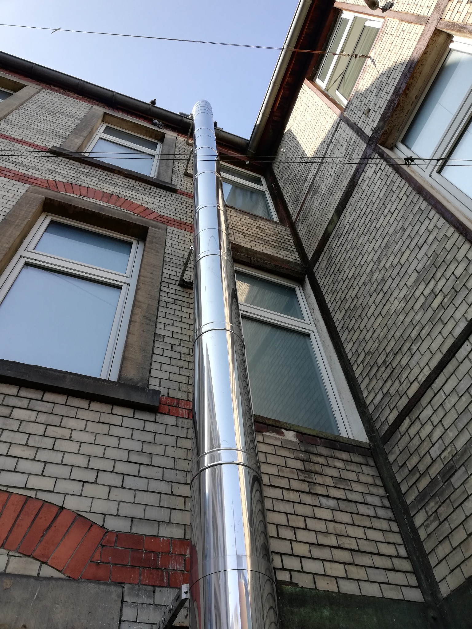 Zewnętrzny, wysoki, metalowy przewód spalinowy, zamontowany na ścianie budynku z cegły, widoczny z dołu pod kątem.