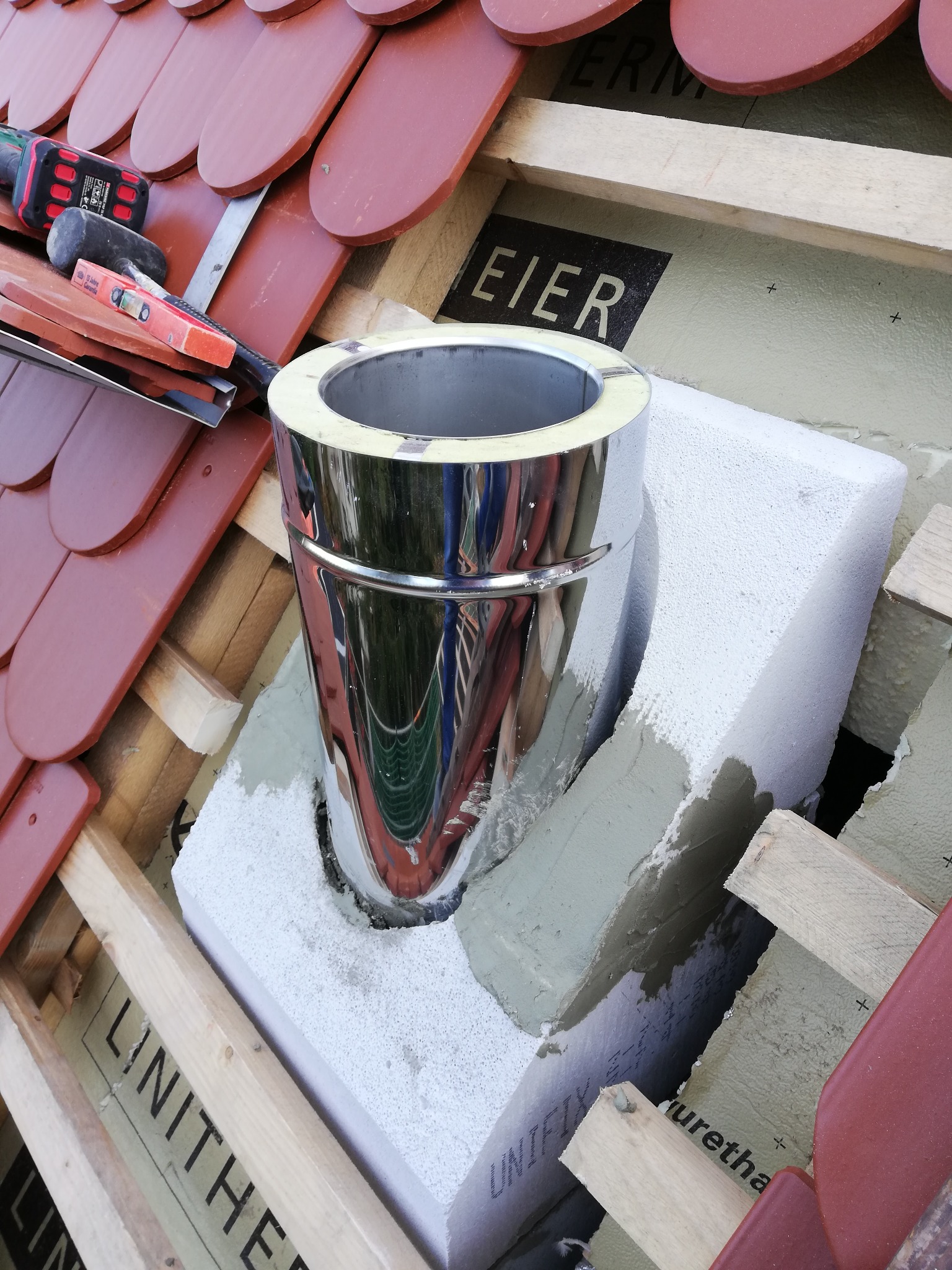 Montaż komina systemowego: chromowany wkład kominowy osadzony w bloczku izolacyjnym na tle drewnianej konstrukcji dachu i czerwonej dachówki, widoczne narzędzia pracy.