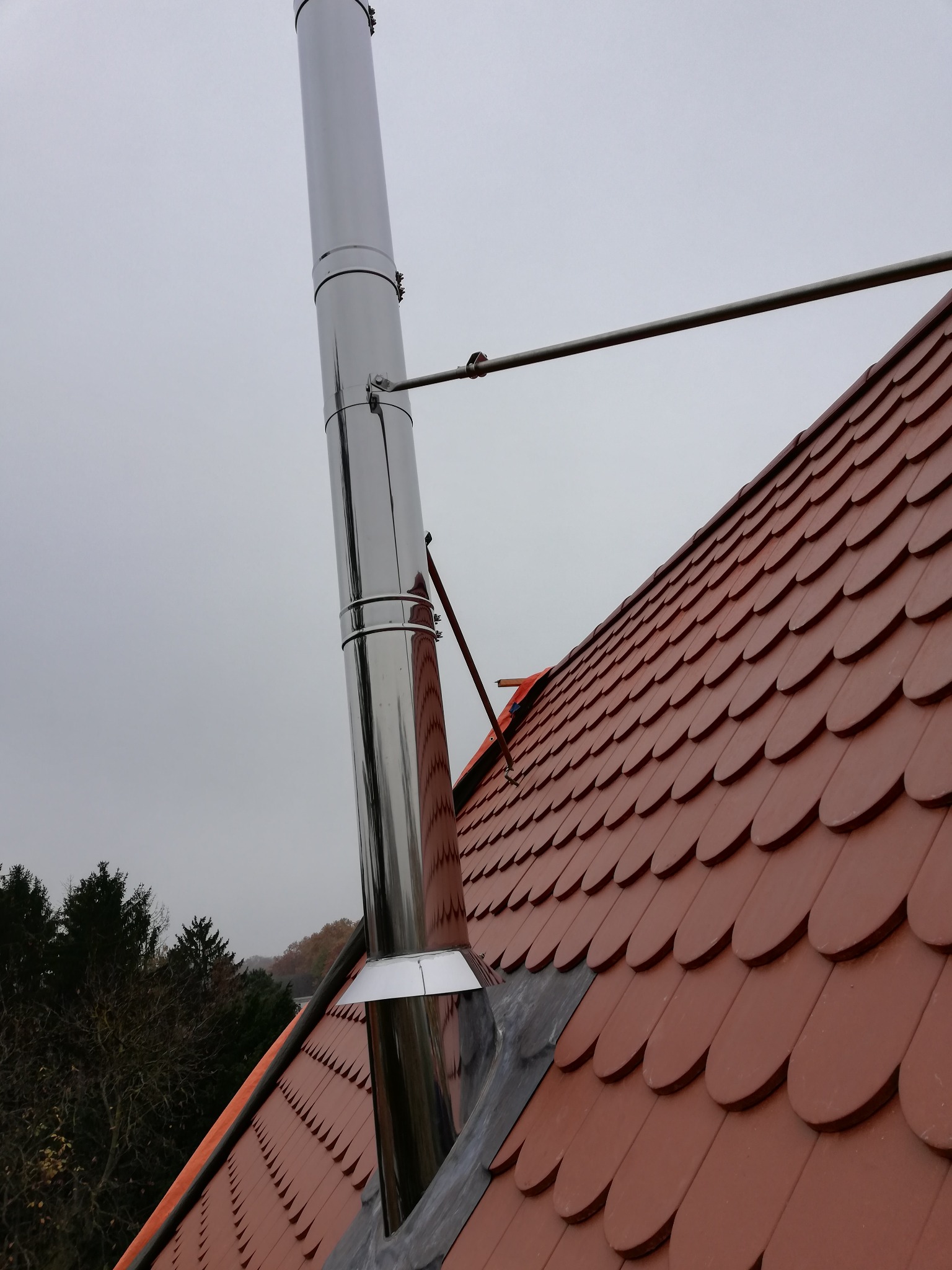 Montaż stalowego przewodu kominowego na dachu z dachówki ceramicznej w kolorze czerwonym, widoczne mocowania i uszczelnienie przy podstawie komina, pochmurne niebo w tle.