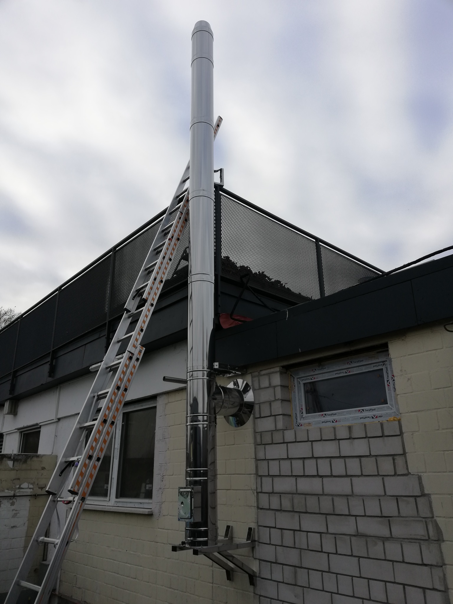 Zewnętrzny, metalowy przewód spalinowy zamontowany na ścianie budynku z cegły, wsparty na metalowych wspornikach, z przystawioną aluminiową drabiną. Widoczne okno z plastikową ramą i metalowa...