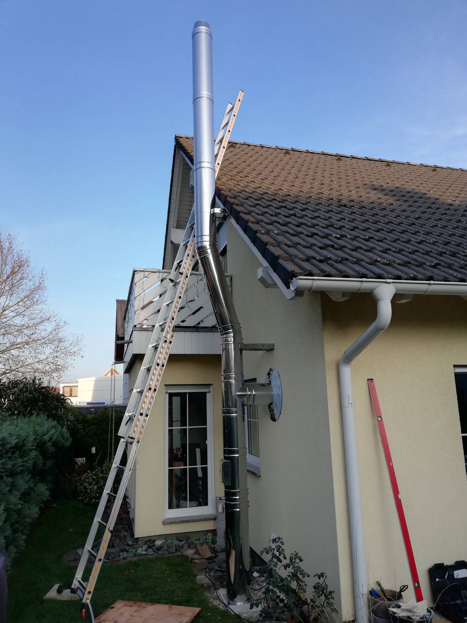 Montaż zewnętrznego, stalowego przewodu spalinowego przy ścianie domu jednorodzinnego. Widoczna drabina oparta o dach i ścianę budynku.