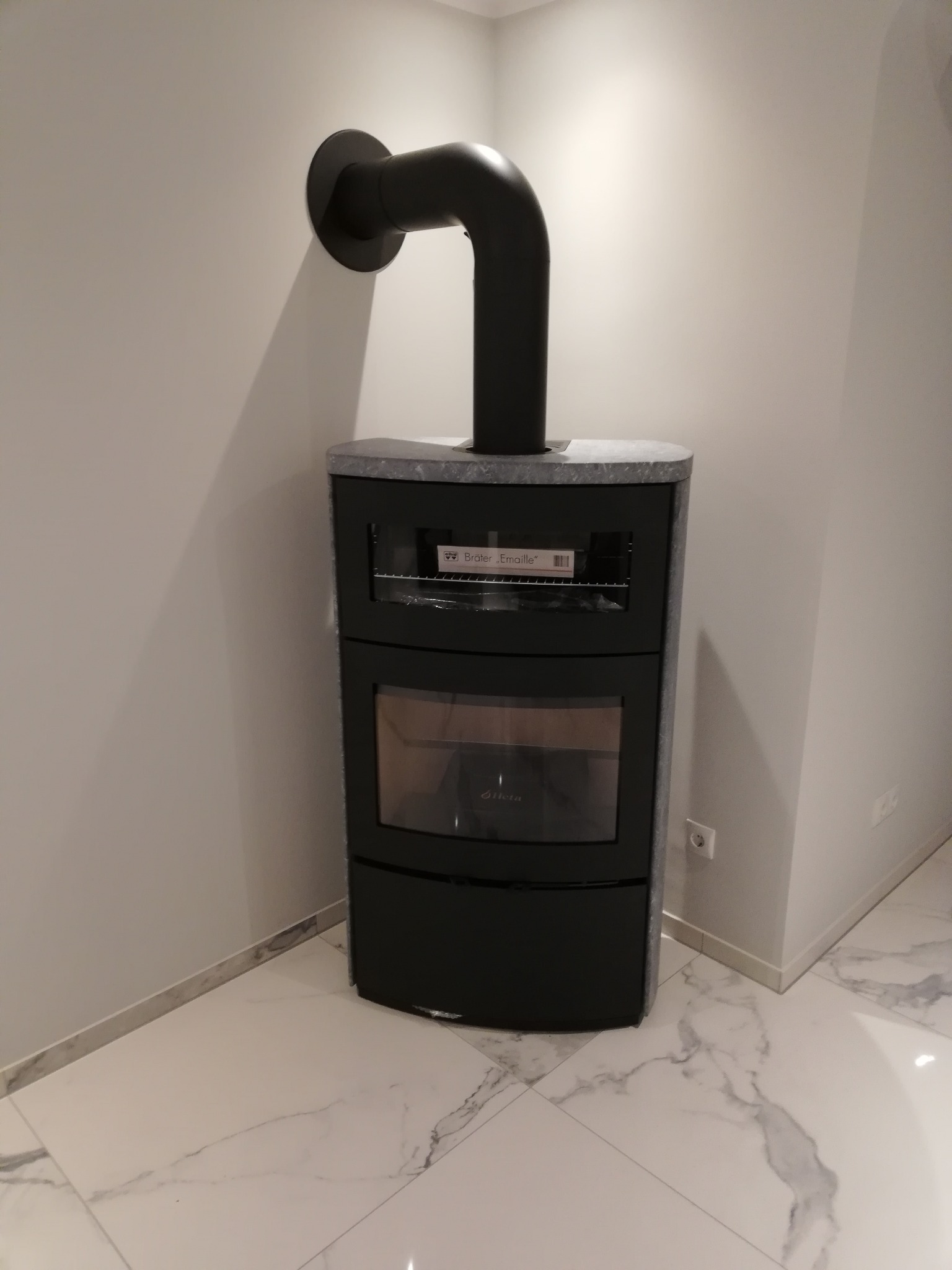 Czarny piec wolnostojący z szarym kamiennym blatem i podłączeniem do komina, ustawiony w rogu pomieszczenia z jasnymi ścianami i marmurową podłogą.