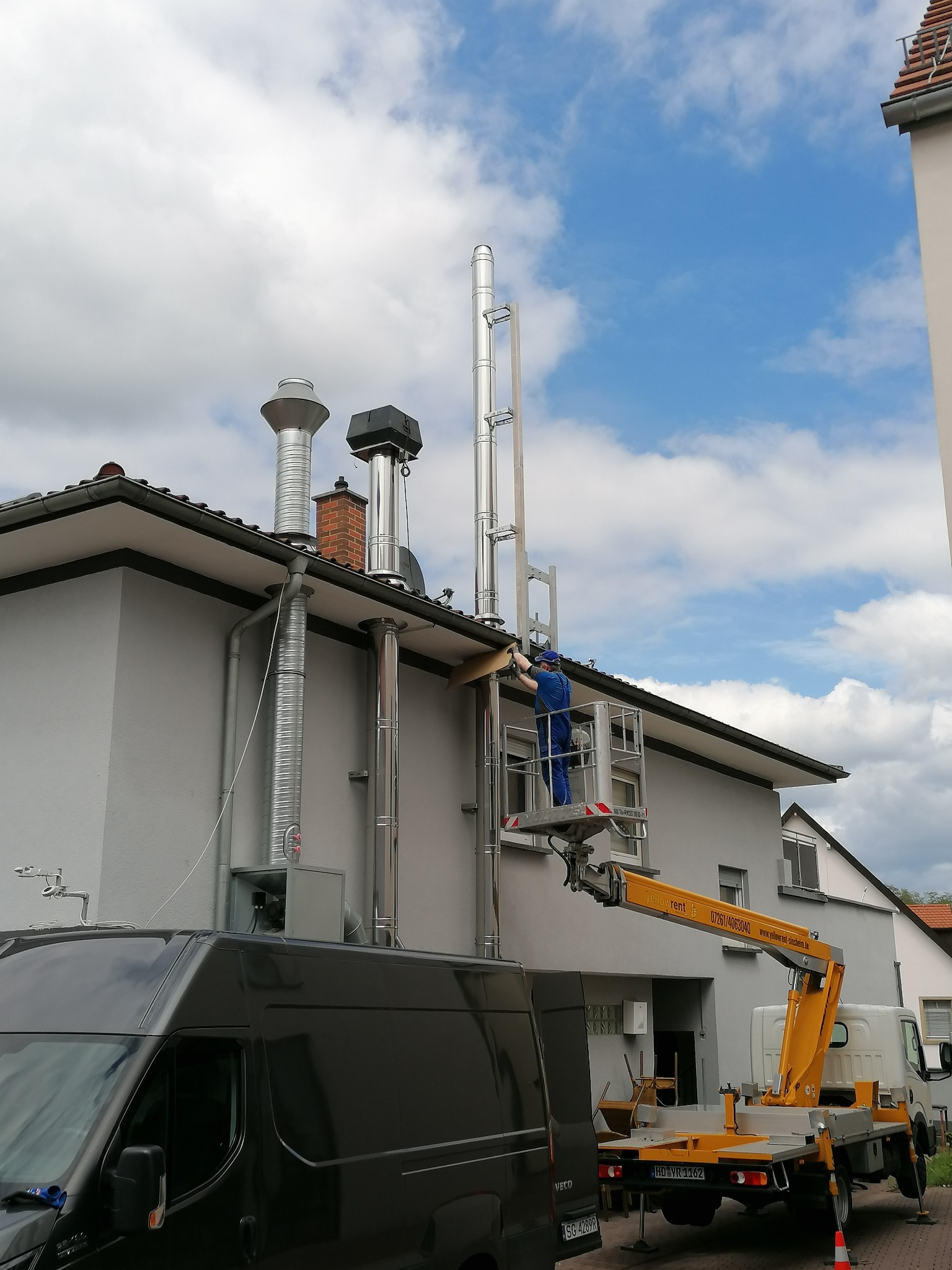 Pracownik w niebieskim kombinezonie na podnośniku koszowym montuje system odprowadzania spalin na dachu szarego domu jednorodzinnego; widoczne elementy kominowe ze stali nierdzewnej i czarny van...