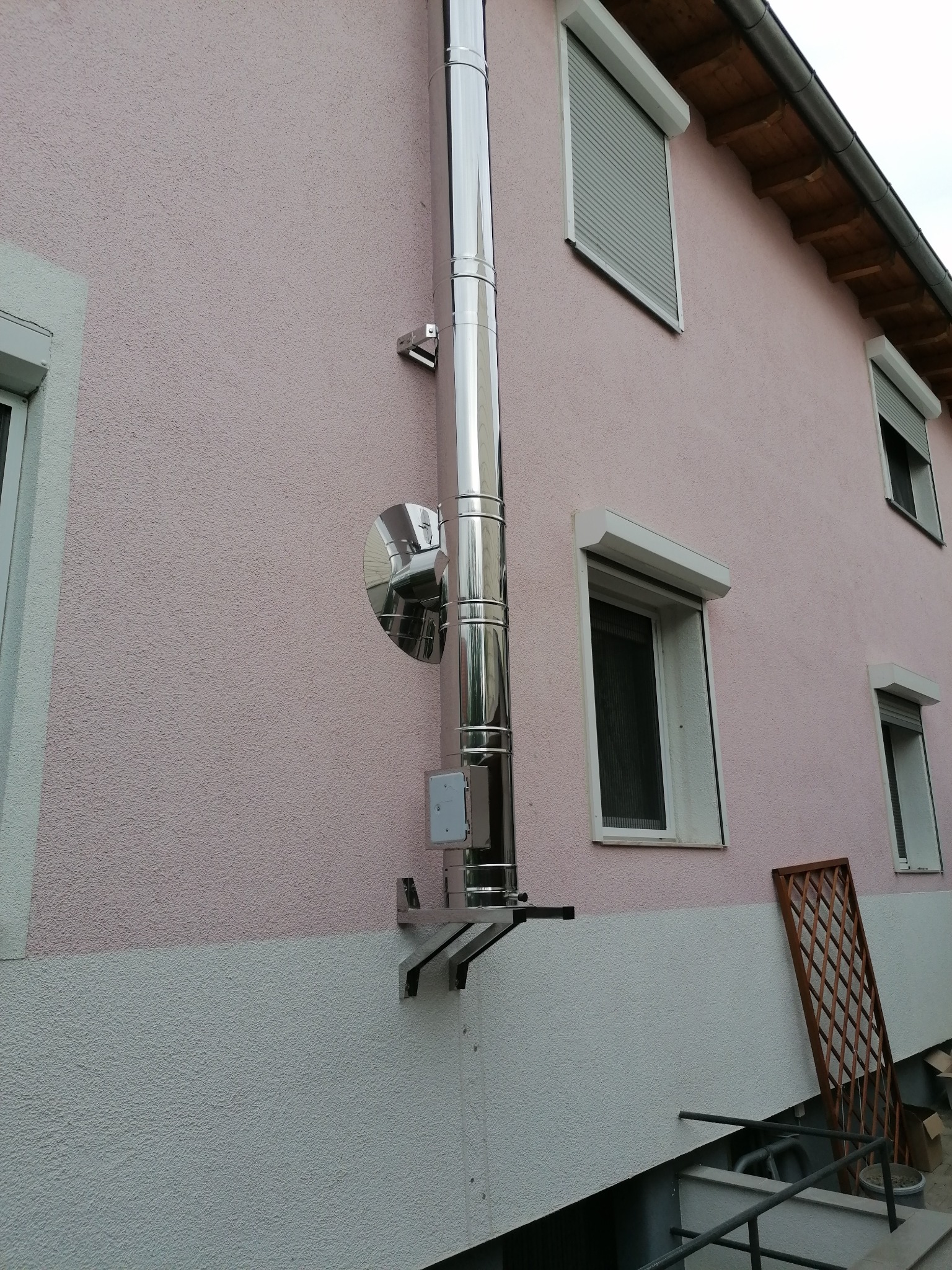 Zewnętrzny, chromowany system odprowadzania spalin, zamontowany na różowej ścianie budynku z widocznymi oknami z roletami.