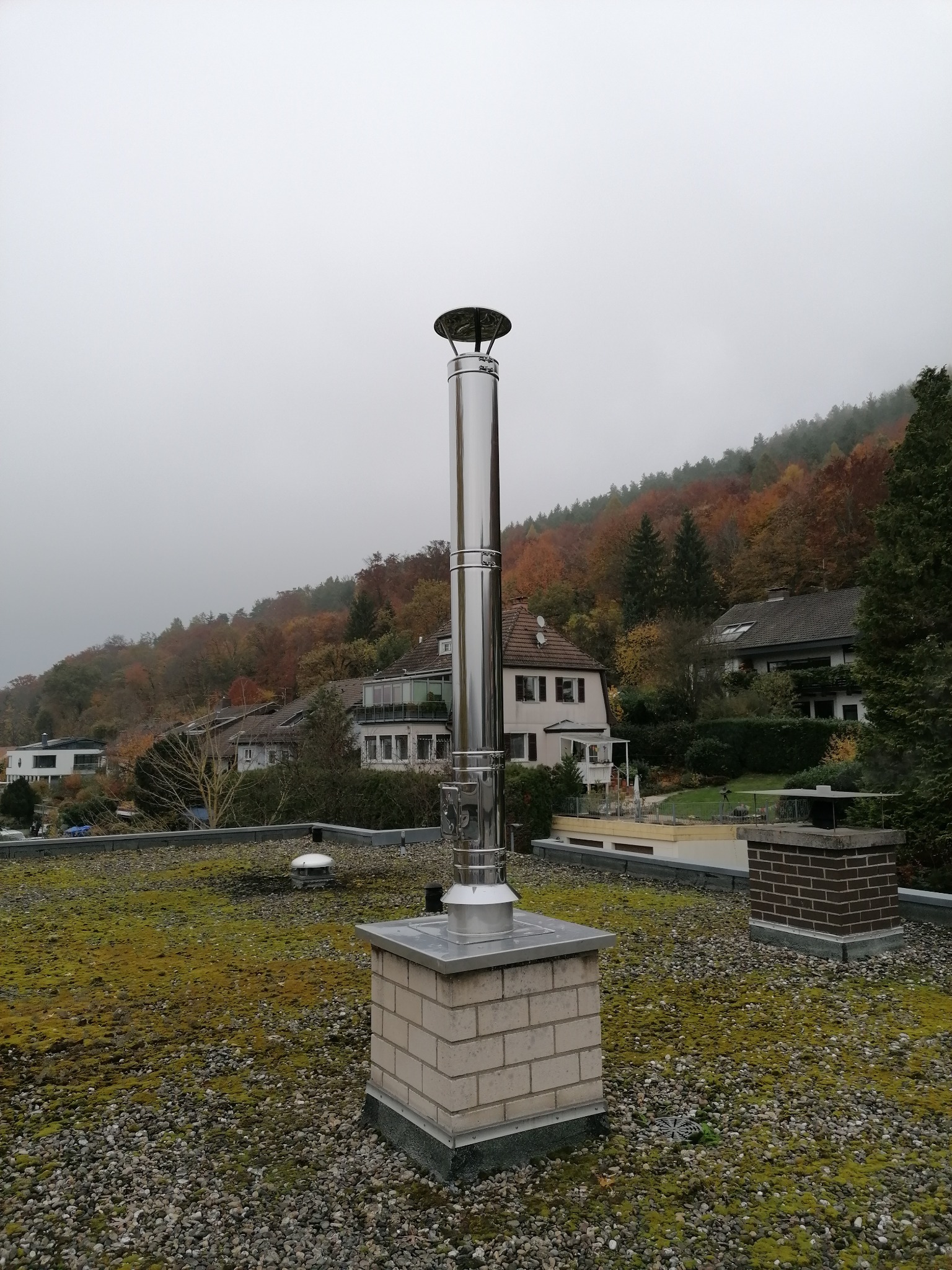 Błyszczący, stalowy komin na ceglanej podstawie, z widokiem na zalesione wzgórze w jesiennych barwach i zabudowania w tle, sfotografowany z dachu pokrytego mchem i drobnym żwirem w pochmurny dzień.