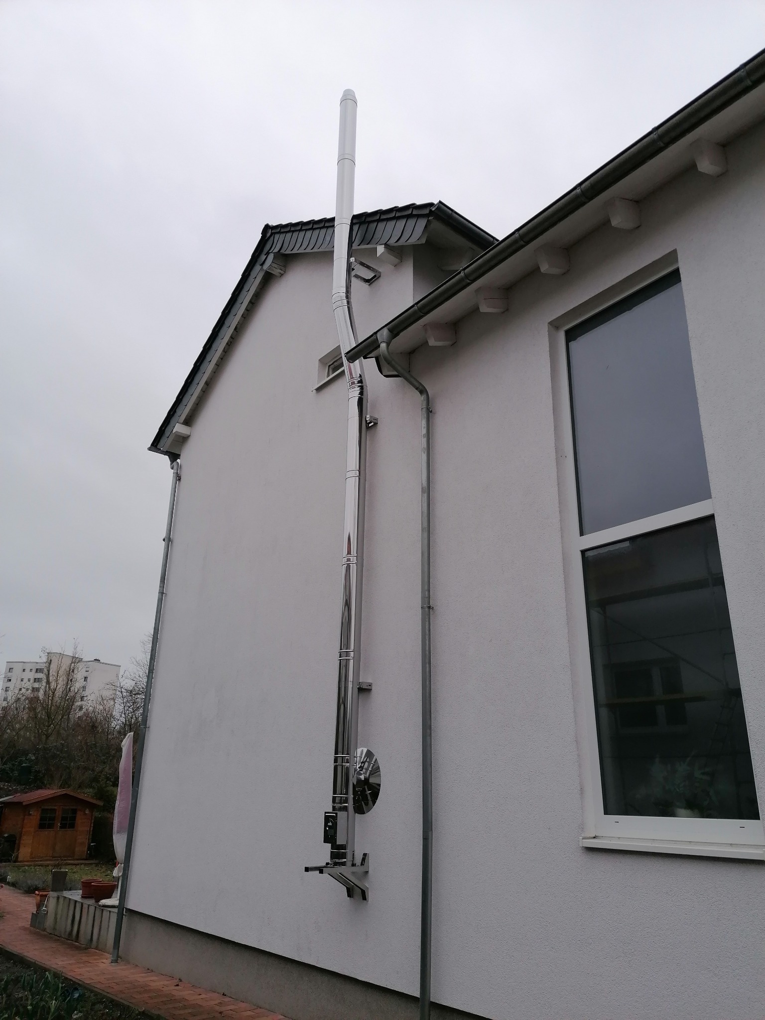 Zewnętrzny, stalowy system odprowadzania spalin zamontowany na ścianie budynku z widocznym oknem i fragmentem dachu pokrytego dachówką. W tle widoczne drzewa i zabudowania.