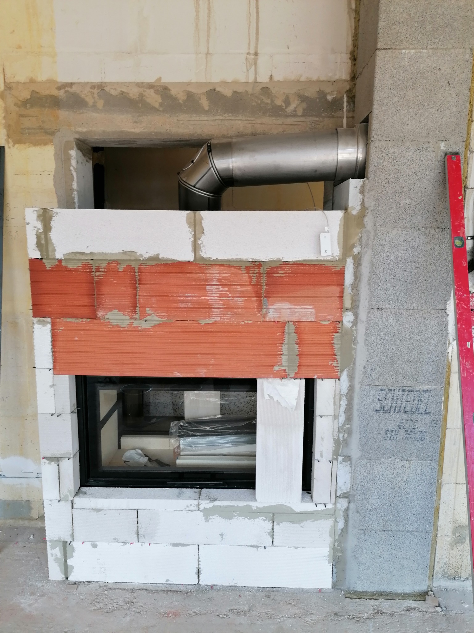 Wnętrze wnęki na wkład kominkowy w trakcie obudowy z białych bloczków i cegły szamotowej, widoczny stalowy przewód kominowy i poziomica.