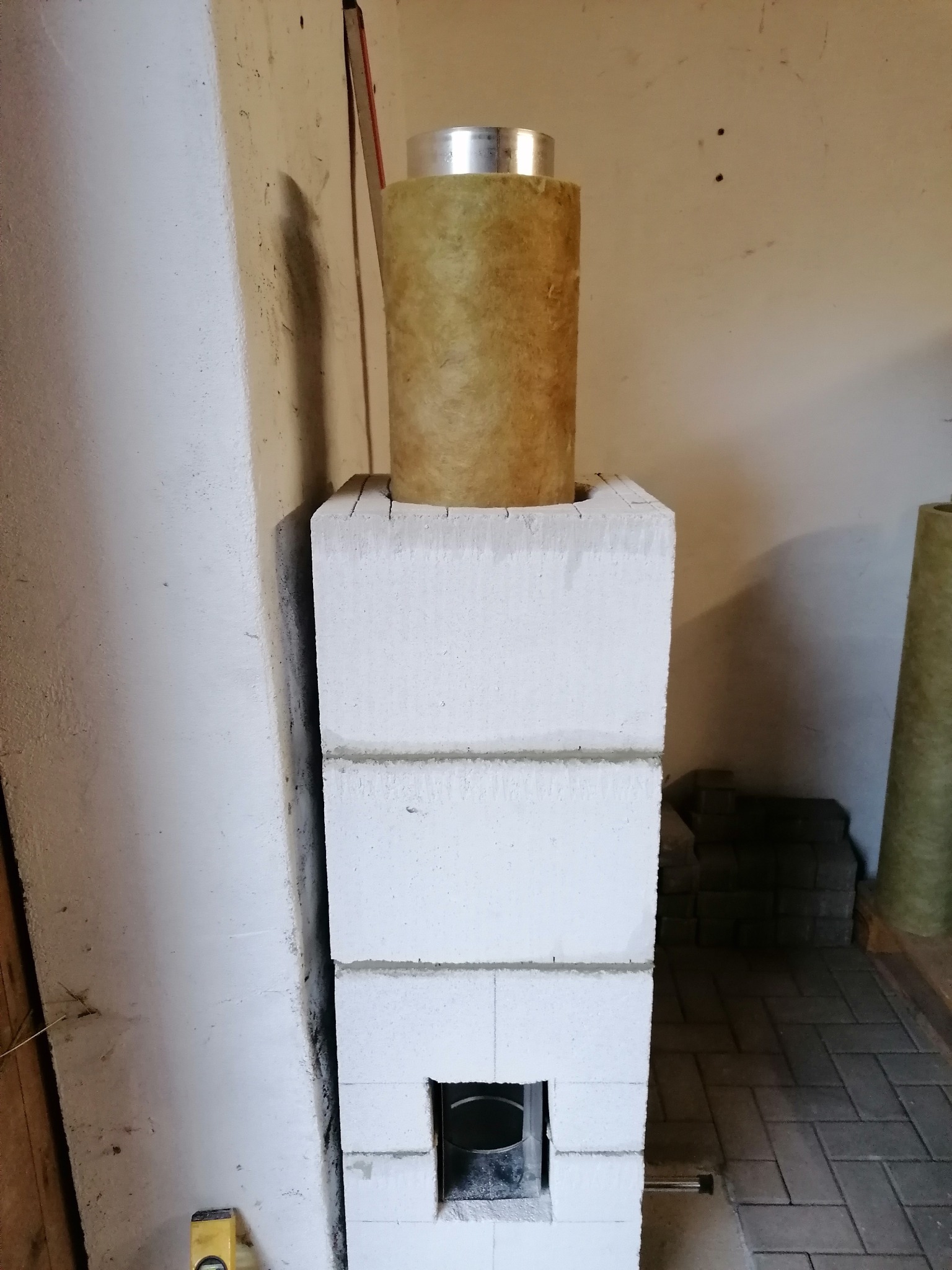 Wznoszenie obudowy z białych bloczków betonu komórkowego dla wkładu grzewczego, z widocznym otworem rewizyjnym i zamontowanym izolowanym przewodem kominowym, na tle surowej ściany w trakcie prac...