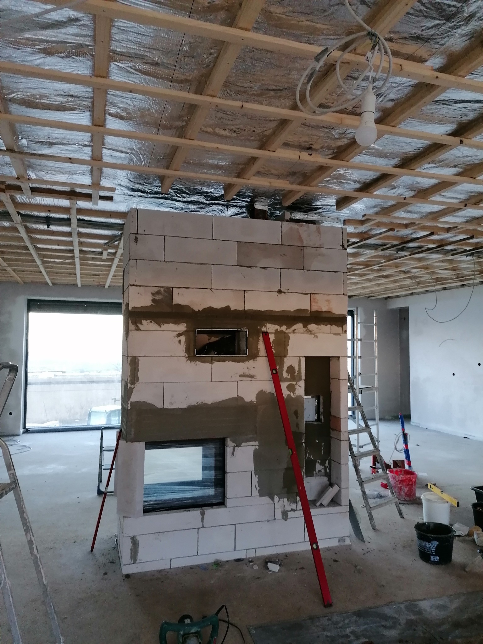 Budowa obudowy wkładu kominkowego z białych bloczków, widoczny poziomica, w tle surowy strop i okno.