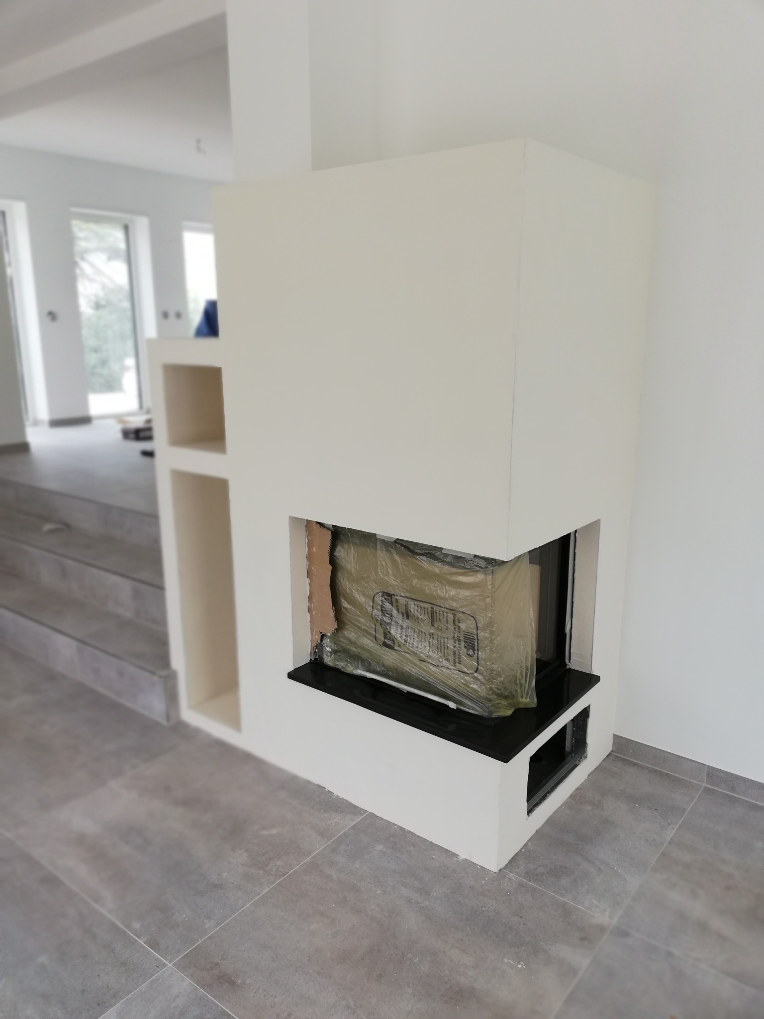 Nowoczesna obudowa kominkowa w trakcie montażu, z widocznym wkładem zabezpieczonym folią, jasne ściany i szare płytki podłogowe.