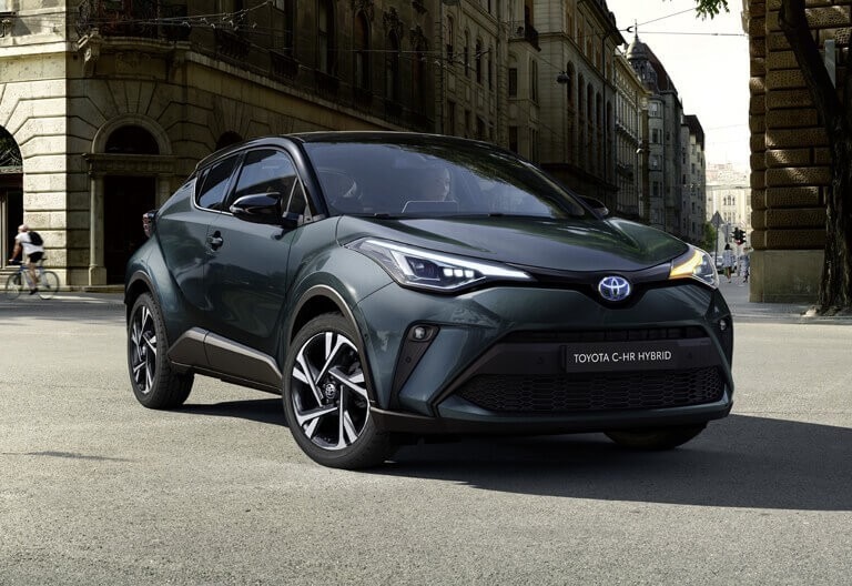 Ciemnozielony samochód Toyota C-HR Hybrid zaparkowany na tle miejskiej zabudowy z pieszym na rowerze w tle.