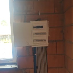 ElectroPointSystem - Biała skrzynka elektryczna z otwartymi drzwiczkami, zamontowana na ceglanej ścianie w surowym stanie, z widocznymi przewodami w peszlach.