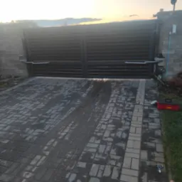 Nowoczesna, przesuwna brama wjazdowa z automatyką, zamontowana na posesji z kostki brukowej. Widoczny napęd bramy i skrzynka narzędziowa. Tło: zachodzące słońce.