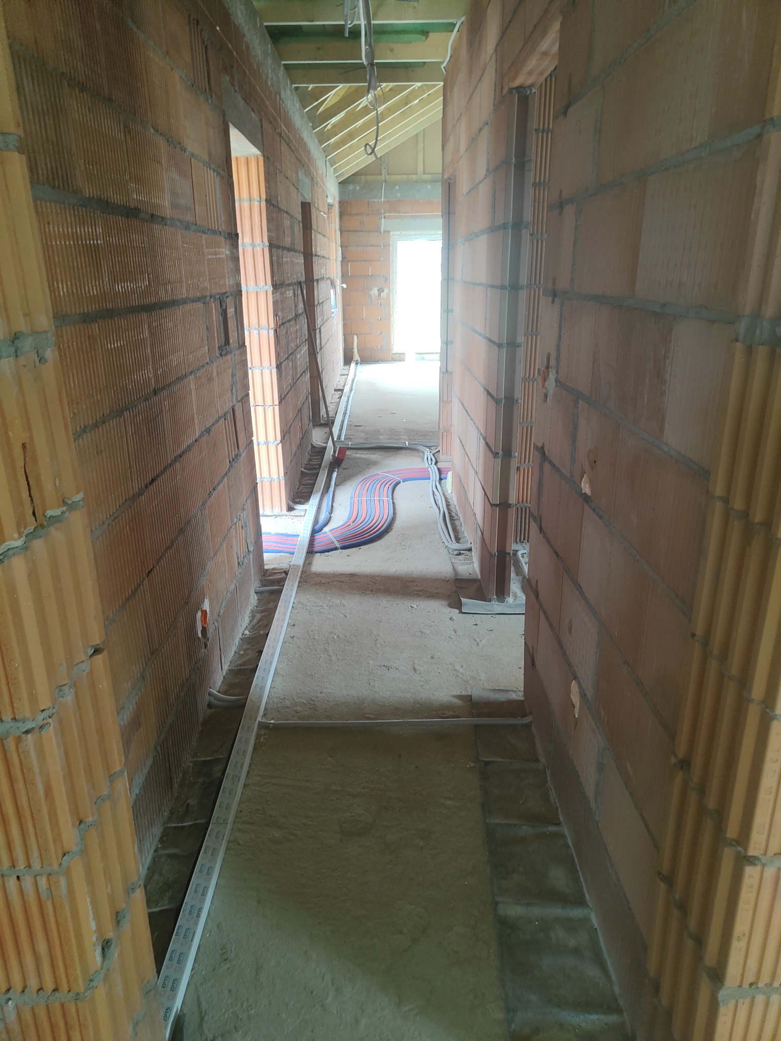 Wnętrze surowego budynku z widoczną instalacją elektryczną w korytarzu, poprowadzoną wzdłuż ściany. Budynek w trakcie budowy, ściany z cegły dziurawki.