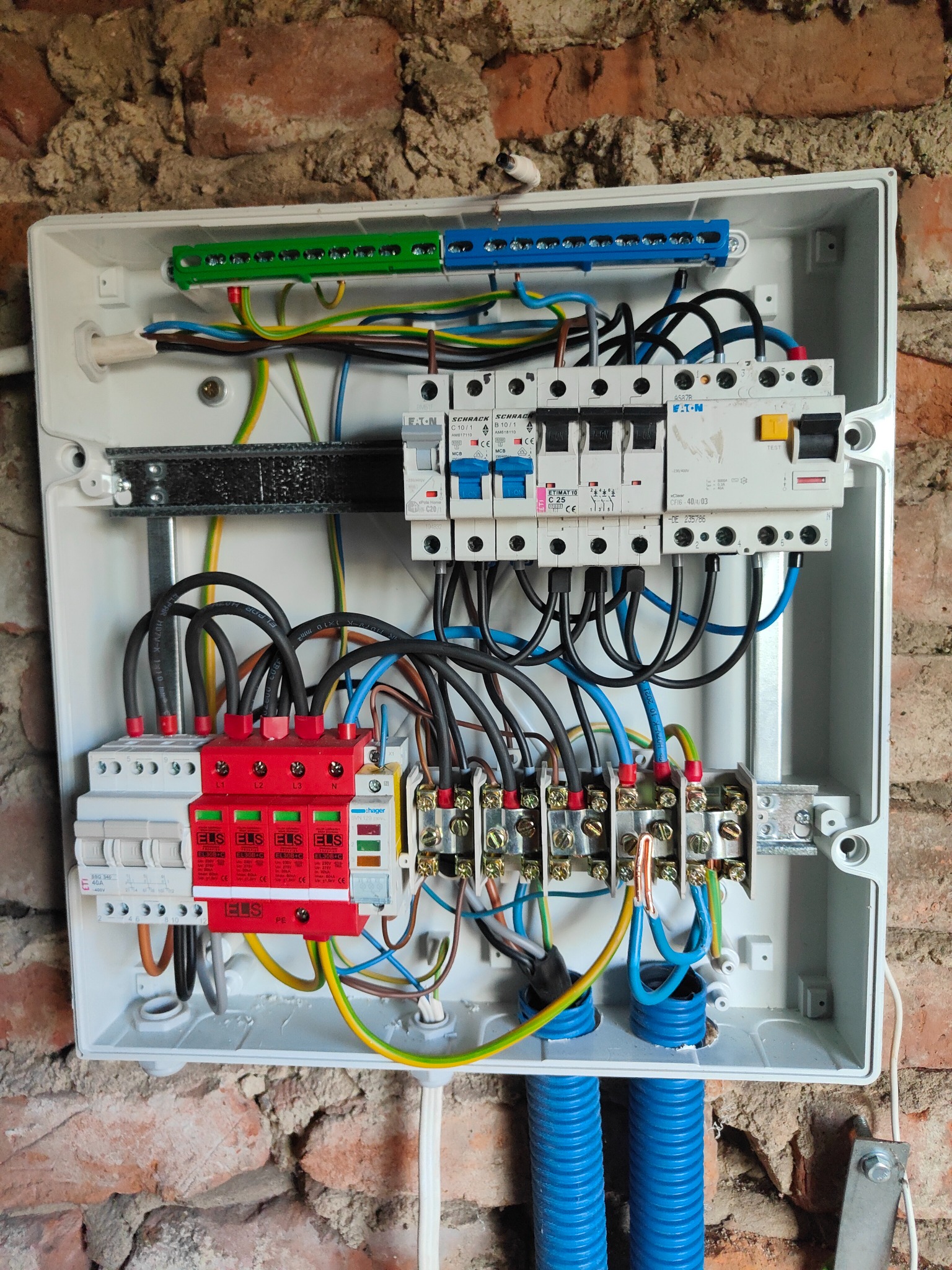 Rozdzielnica elektryczna z zainstalowanymi zabezpieczeniami przepięciowymi ELS, wyłącznikami Eaton i Schrack, zamontowana na ceglanej ścianie, z widocznymi kablami i elementami instalacji.