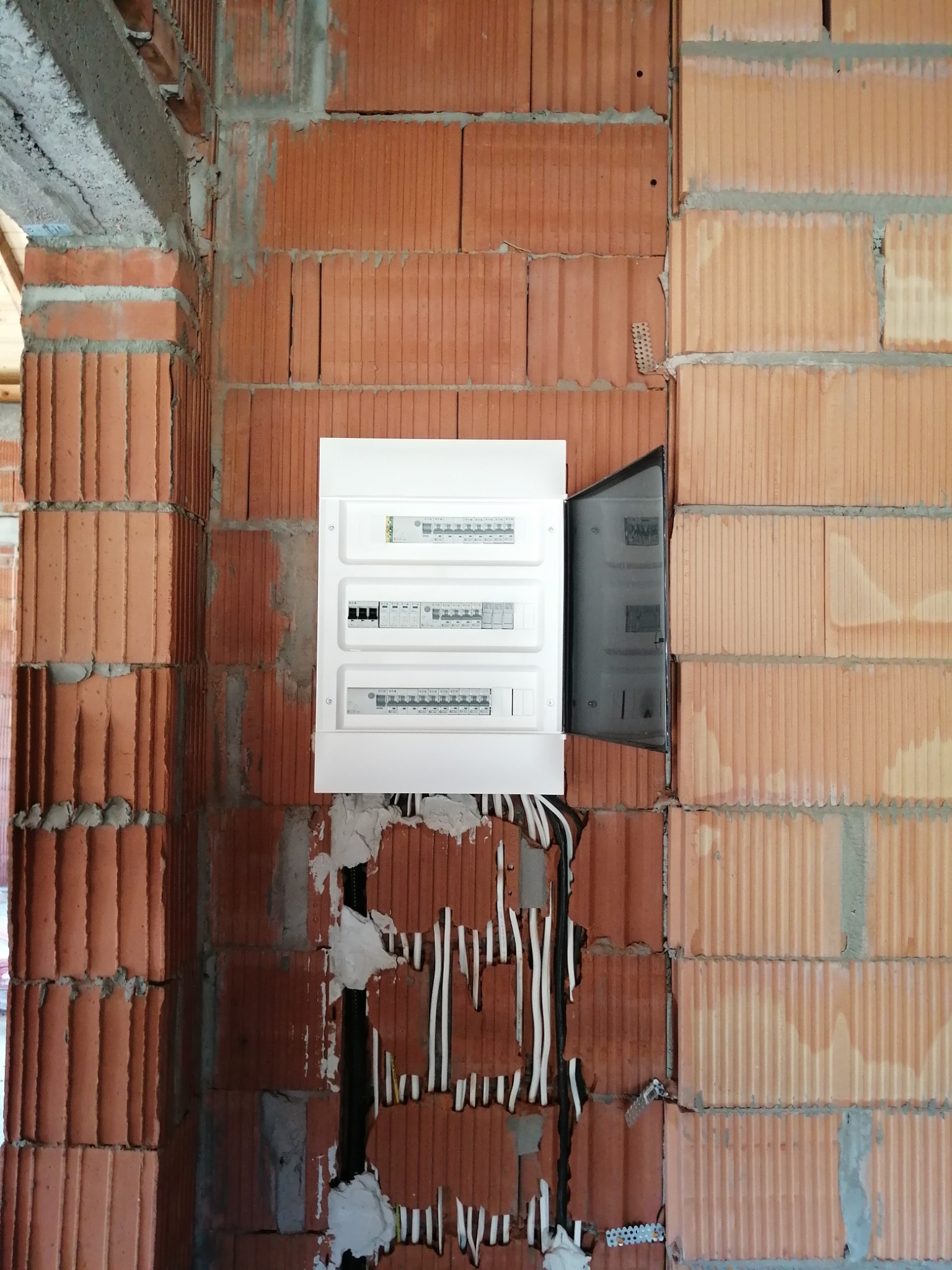 Rozdzielnica elektryczna natynkowa, biała, z otwartymi drzwiczkami, zamontowana na ścianie z czerwonej cegły dziurawki w surowym stanie.