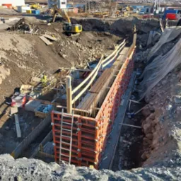 Budowa fundamentów pod mury oporowe: widok na wykop z deskowaniem, zbrojeniem i pracującym robotnikiem w oddali, na tle krajobrazu z górami i dźwigiem budowlanym.