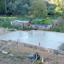 Świeżo wylana płyta fundamentowa z wystającymi prętami zbrojeniowymi i rurami instalacyjnymi, otoczona drewnianym szalunkiem, na tle działki z drzewami i stertami materiałów budowlanych.