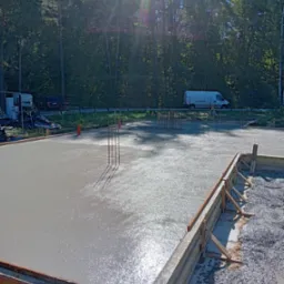 Świeżo wylana, błyszcząca w słońcu posadzka betonowa z widocznym zbrojeniem i drewnianym szalunkiem, w tle las i biały samochód dostawczy.