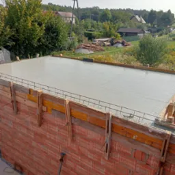 Świeżo wylany, gładki strop betonowy w drewnianym szalunku, z widocznym zbrojeniem, na tle murowanej ściany z czerwonej cegły dziurawki, z podporą stropową i elementami zbrojenia na podłożu. W tle...