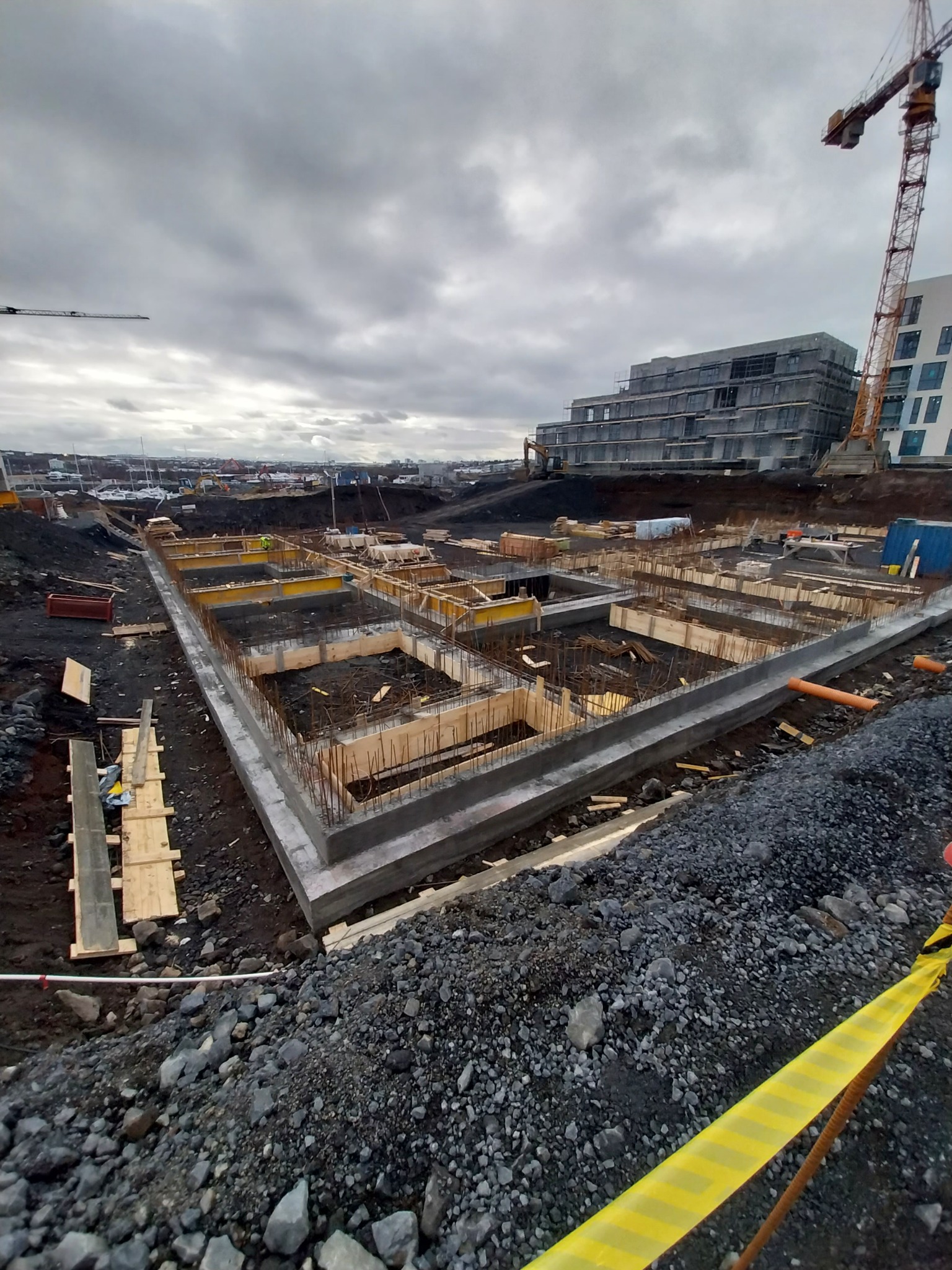 Wykop pod fundamenty z deskowaniem i zbrojeniem, widoczne rury kanalizacyjne, w tle budynek w budowie i żuraw, pochmurne niebo.