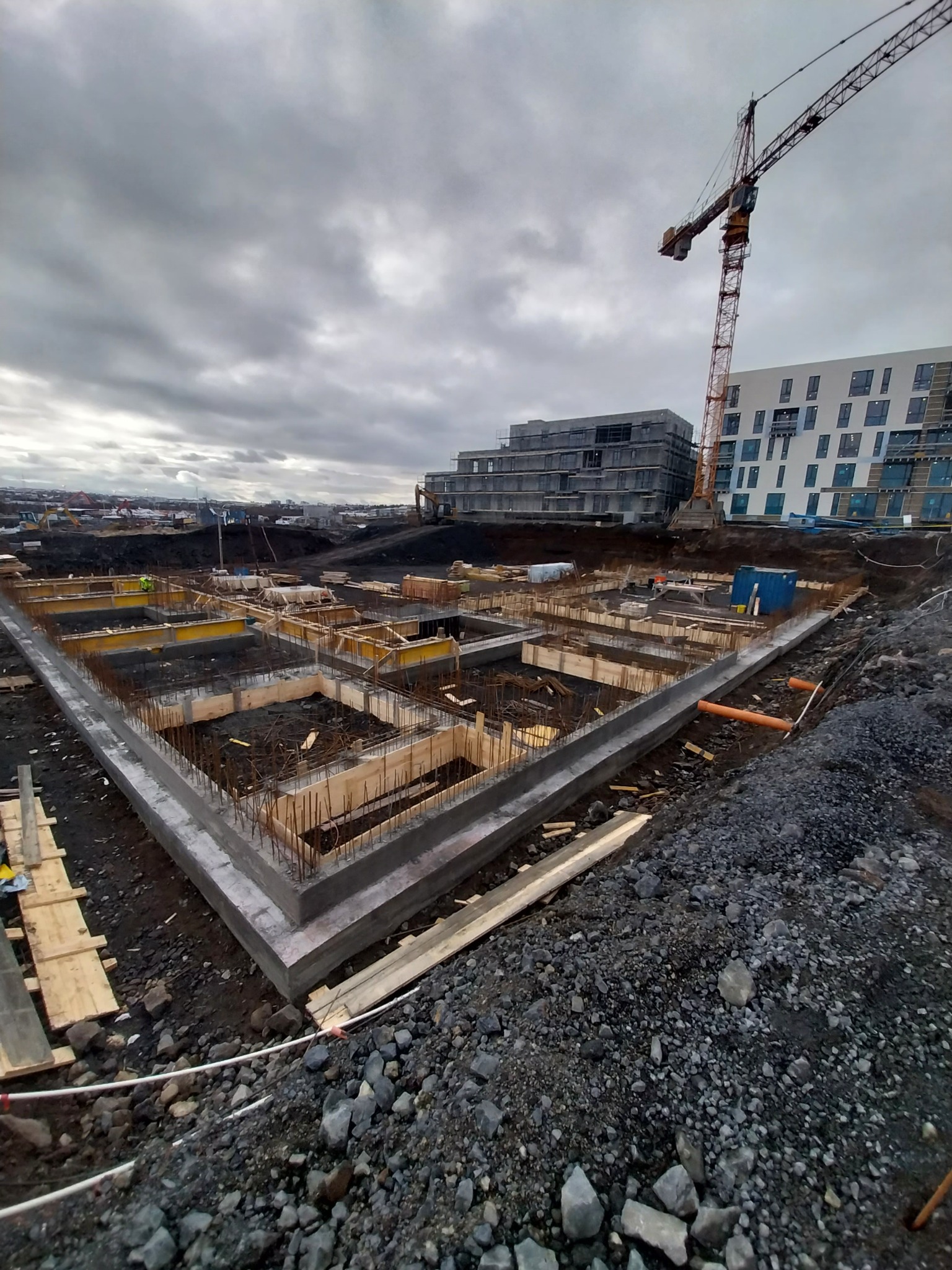 Zalany betonowy fundament z deskowaniem i wystającą stalą zbrojeniową na placu budowy, w tle budynki w trakcie realizacji i żuraw wieżowy, pochmurne niebo.