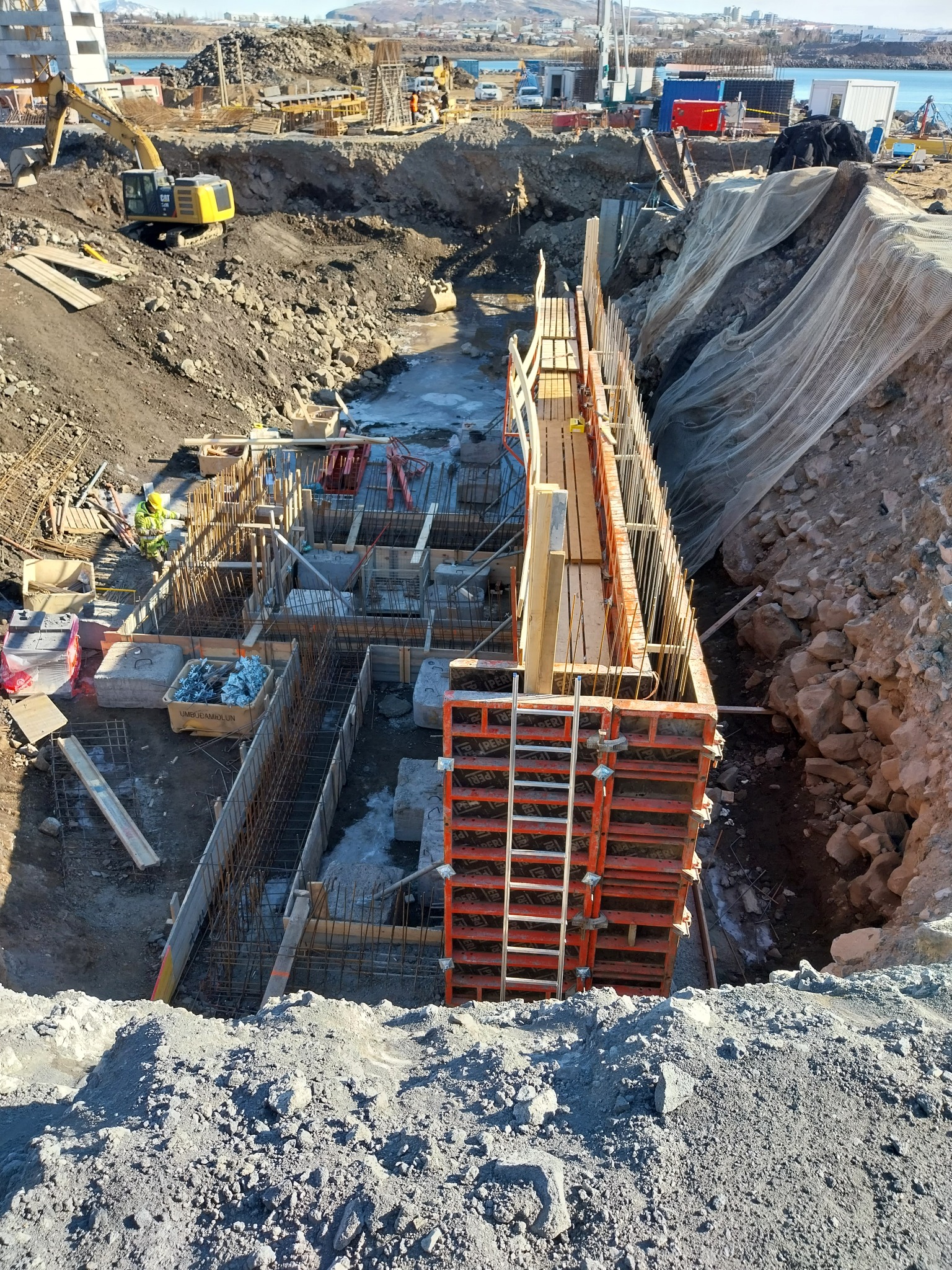 Budowa fundamentów z widocznym szalunkiem, zbrojeniem i pracownikiem w odblaskowej kamizelce na tle wykopu i krajobrazu miejskiego.