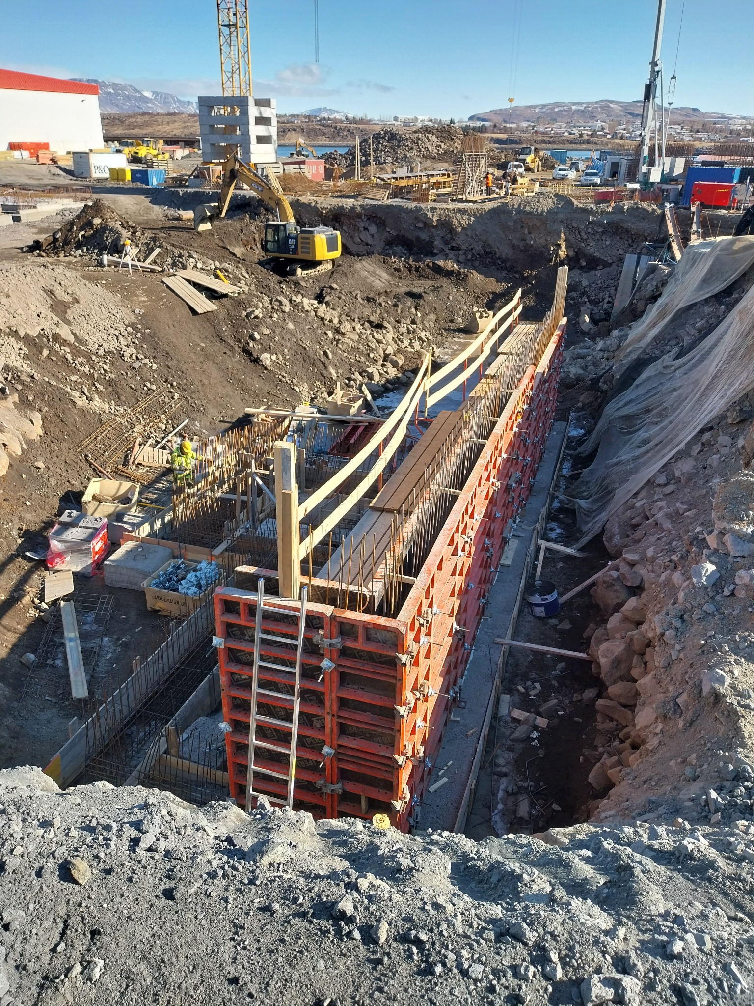 Budowa fundamentów pod mury oporowe: widok na wykop z deskowaniem, zbrojeniem i pracującym robotnikiem w oddali, na tle krajobrazu z górami i dźwigiem budowlanym.