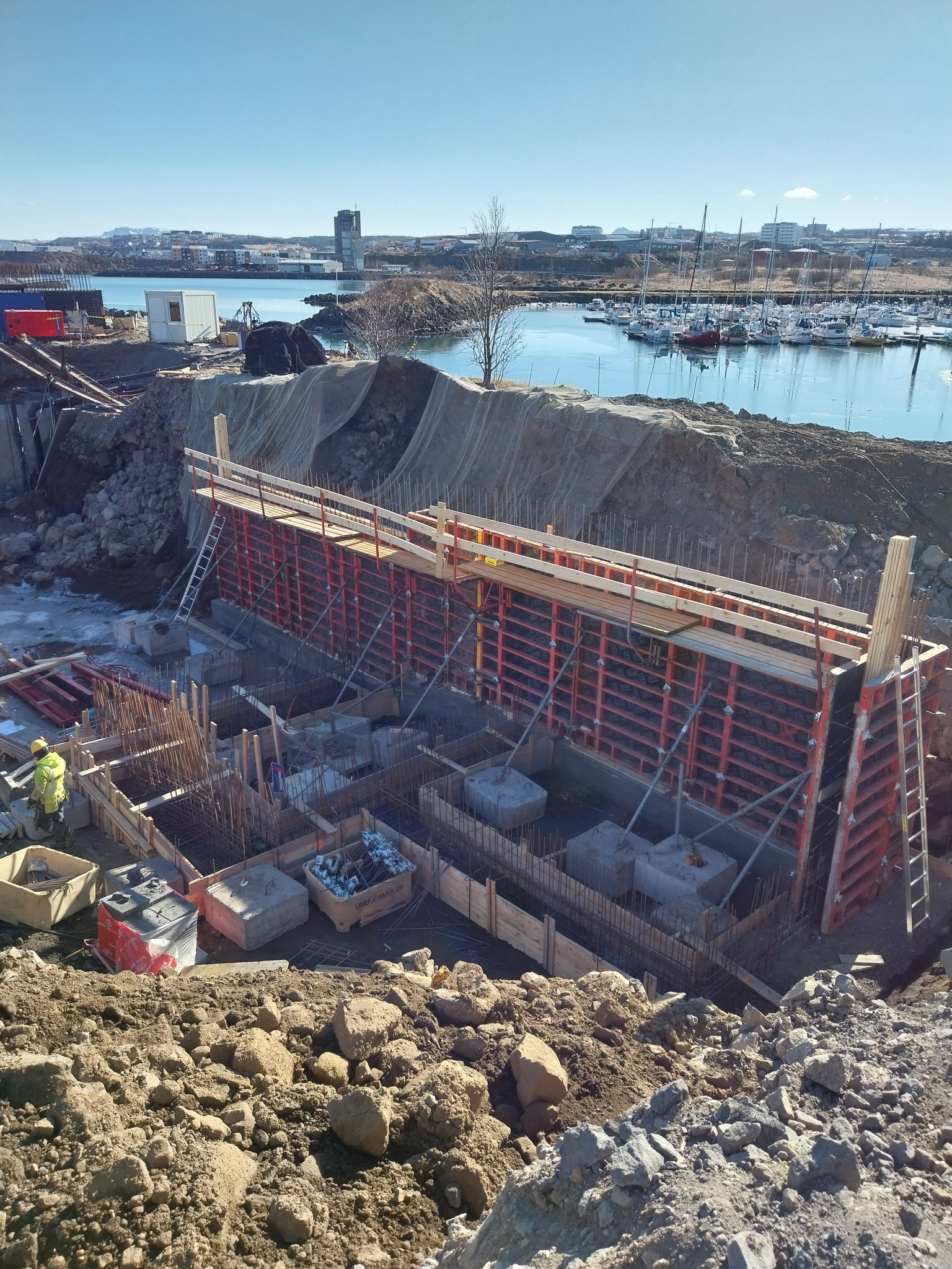 Budowa fundamentów przy nabrzeżu, widoczne zbrojenia, szalunki i betonowe podstawy, w tle marina z jachtami i zabudowa miejska.