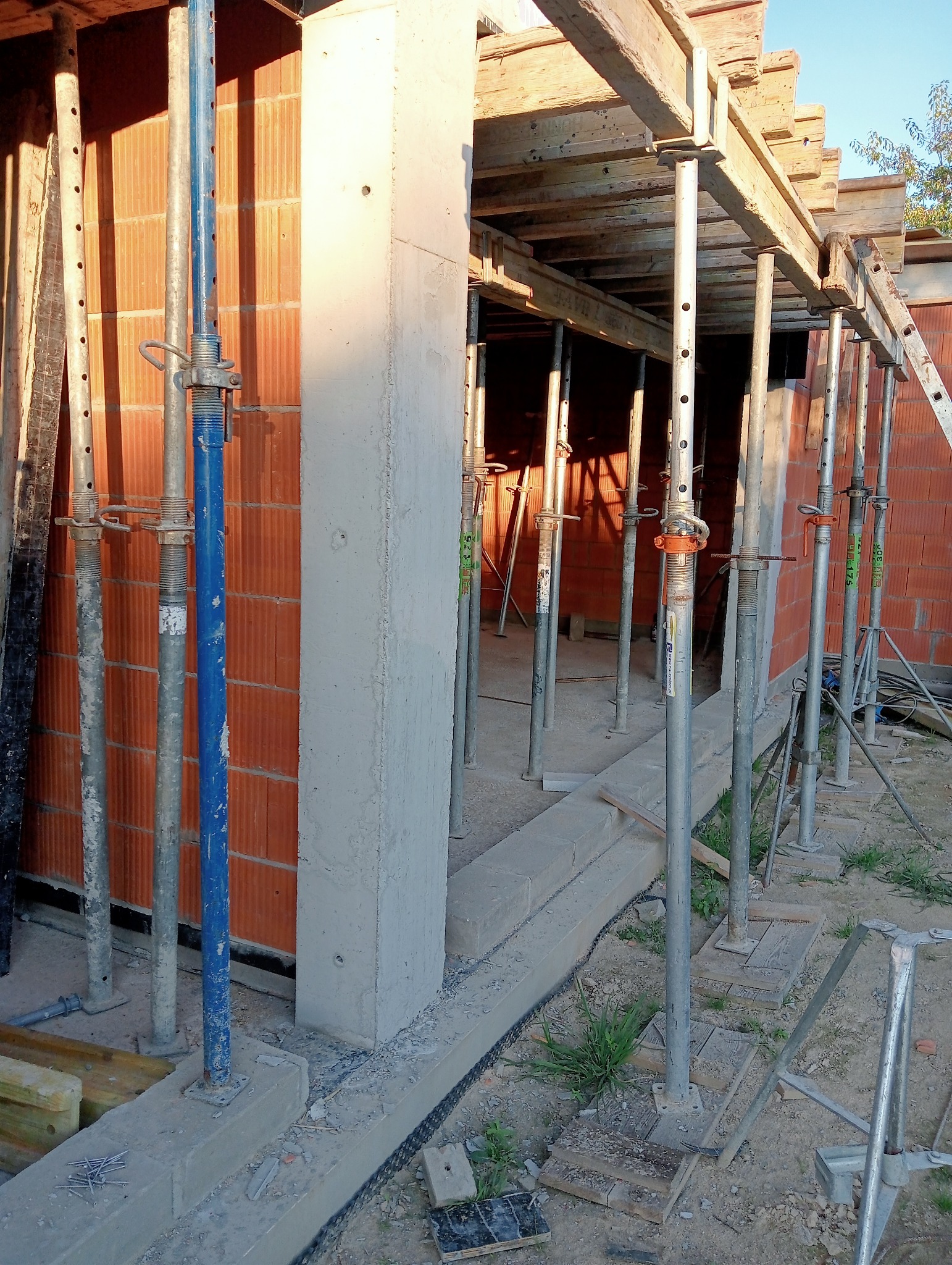 Wznoszenie konstrukcji budynku: widok na betonowy słup, fragment murowanej ściany z pustaków ceramicznych oraz metalowe podpory szalunkowe z drewnianym szalunkiem stropu w trakcie budowy.