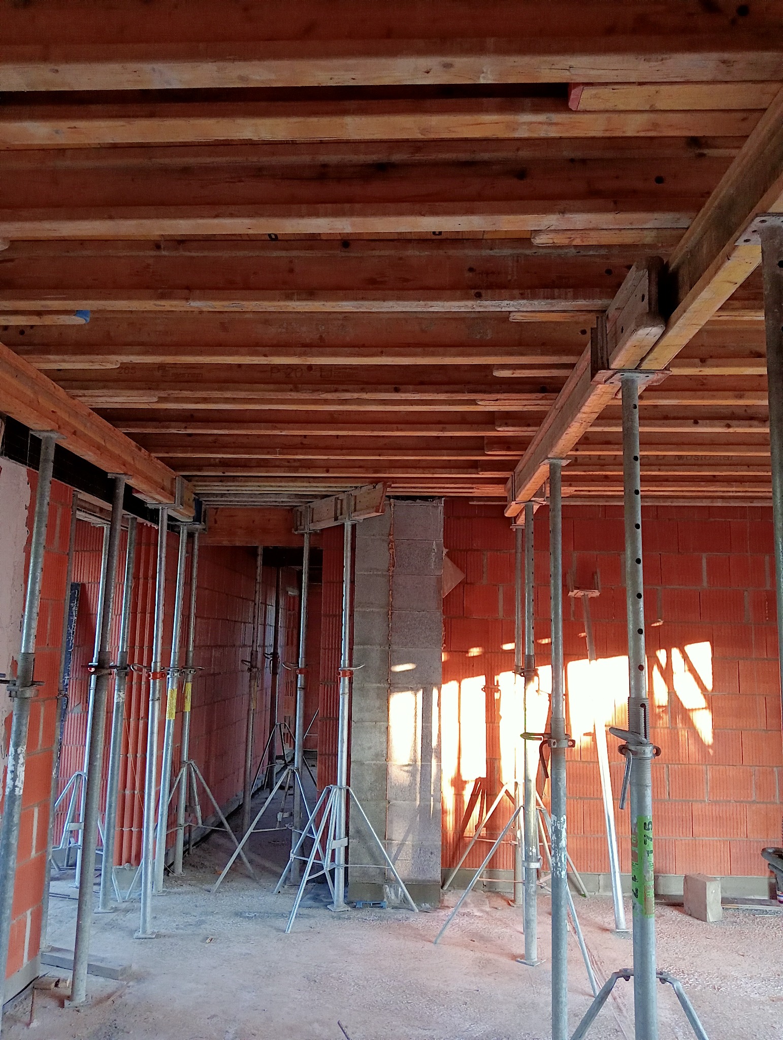 Wnętrze budynku w trakcie budowy, widoczne murowane ściany z czerwonej cegły, betonowy filar, metalowe podpory stropu i drewniane szalunki stropowe.