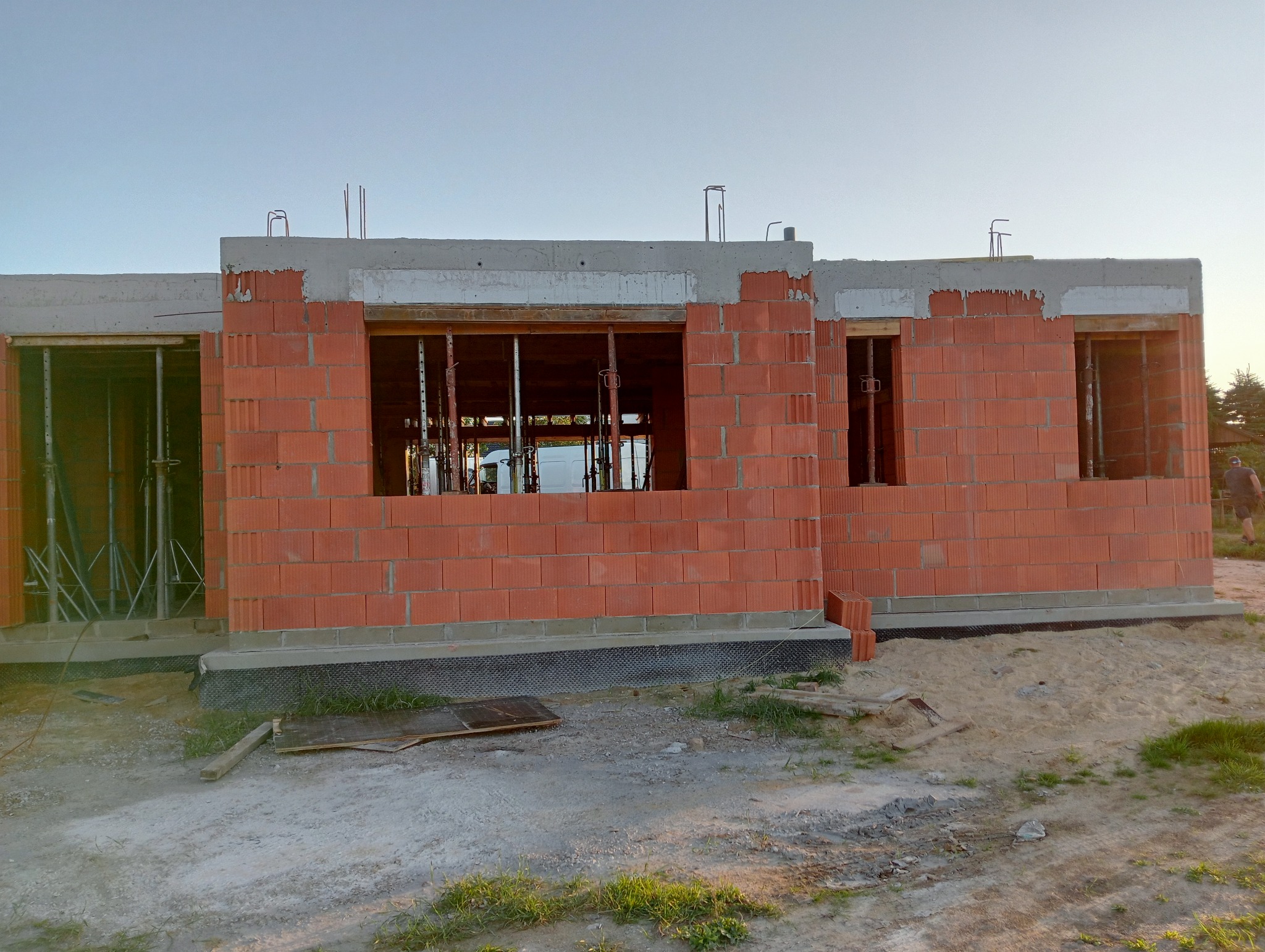 Surowy stan budynku z czerwonej cegły, widoczne otwory okienne i drzwiowe ze wspornikami, fundamenty z izolacją przeciwwilgociową, teren budowy w piasku i trawie.