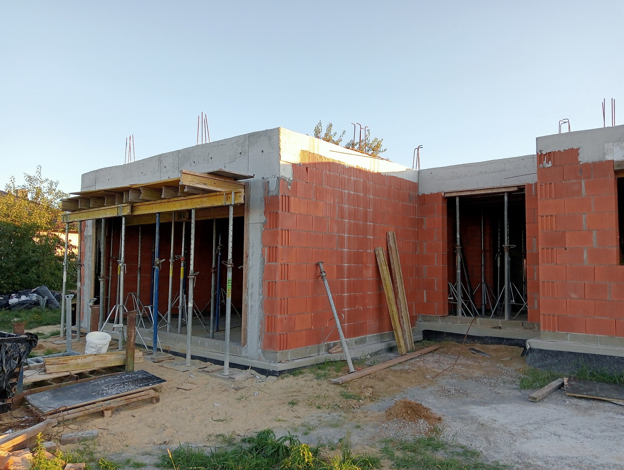 Wznoszenie ścian z czerwonej cegły na budowie domu jednorodzinnego, widoczne podpory szalunkowe, zbrojenie wystające z betonu, oraz elementy izolacji fundamentów.