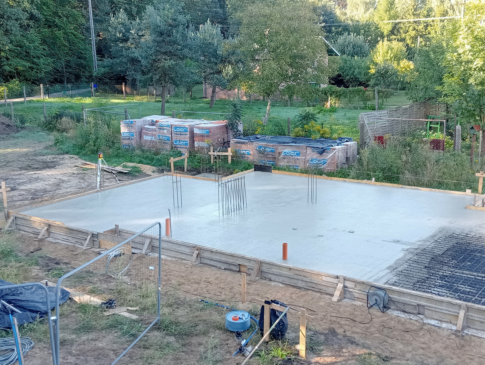 Świeżo wylana płyta fundamentowa z wystającymi prętami zbrojeniowymi i rurami instalacyjnymi, otoczona drewnianym szalunkiem, na tle działki z drzewami i stertami materiałów budowlanych.