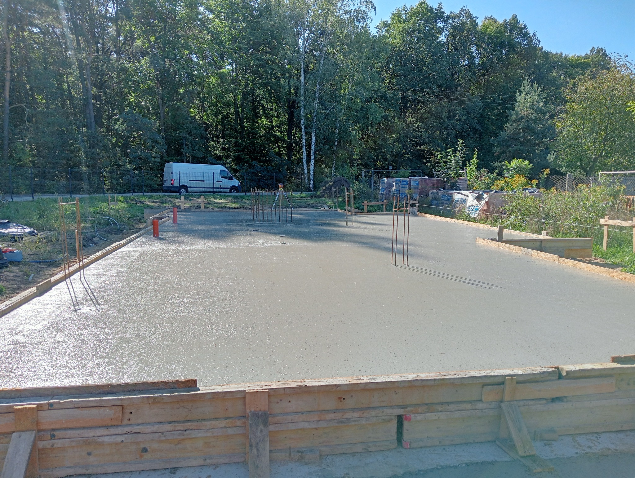 Świeżo wylana, gładka płyta fundamentowa z wystającymi prętami zbrojeniowymi, otoczona drewnianym szalunkiem, na tle zieleni i zaparkowanego białego samochodu dostawczego.