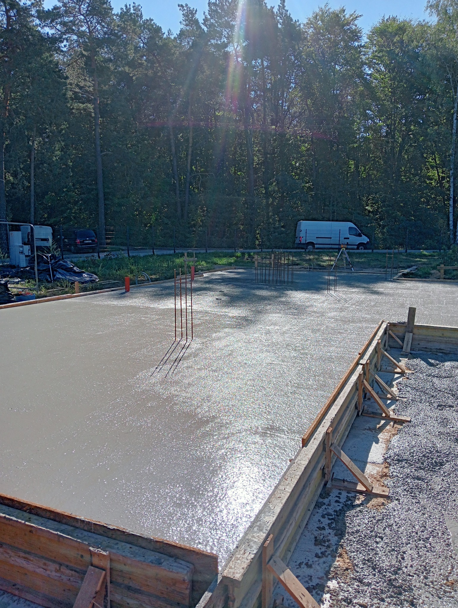 Świeżo wylana, błyszcząca w słońcu posadzka betonowa z widocznym zbrojeniem i drewnianym szalunkiem, w tle las i biały samochód dostawczy.