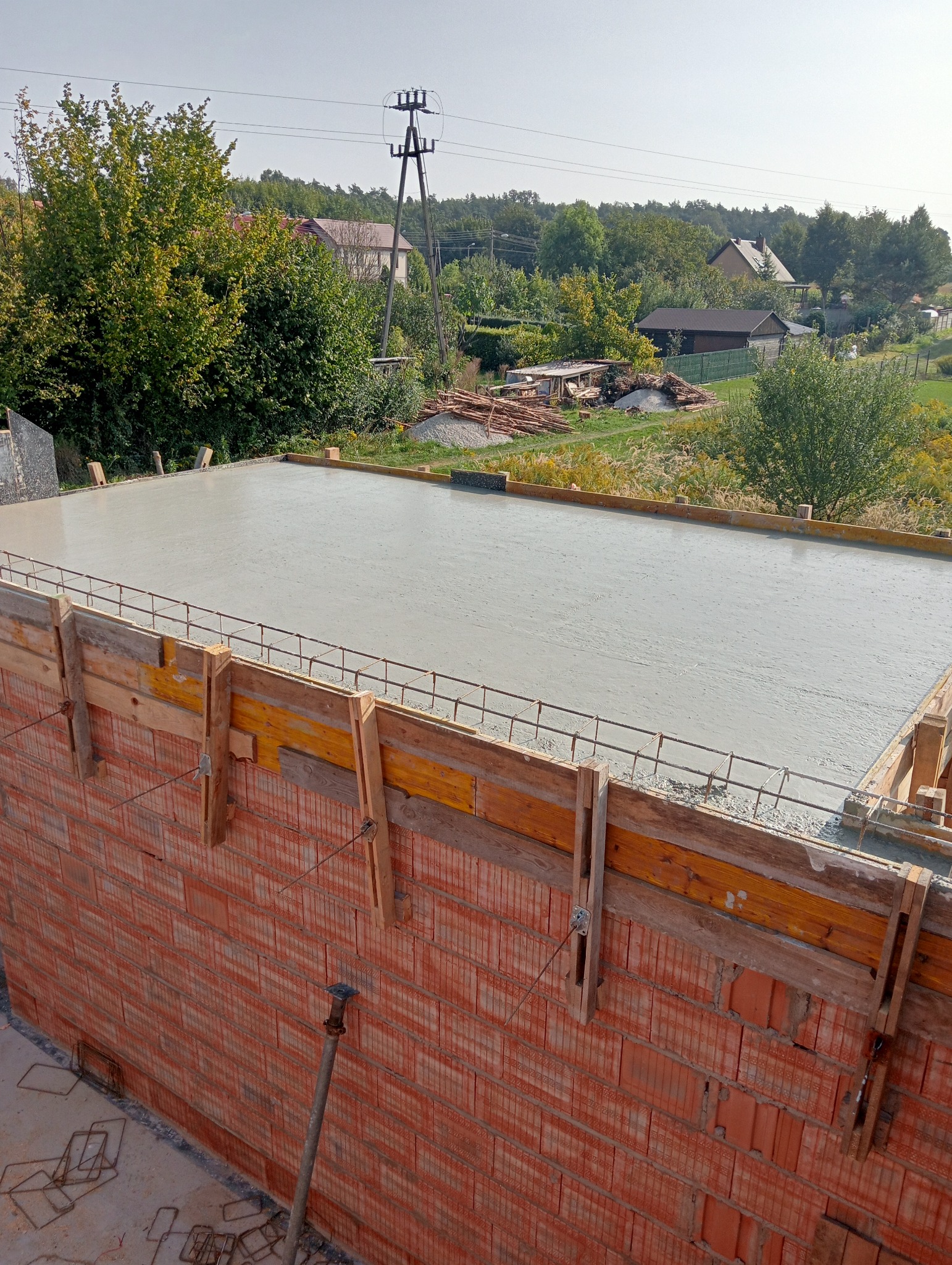 Świeżo wylany, gładki strop betonowy w drewnianym szalunku, z widocznym zbrojeniem, na tle murowanej ściany z czerwonej cegły dziurawki, z podporą stropową i elementami zbrojenia na podłożu. W tle...